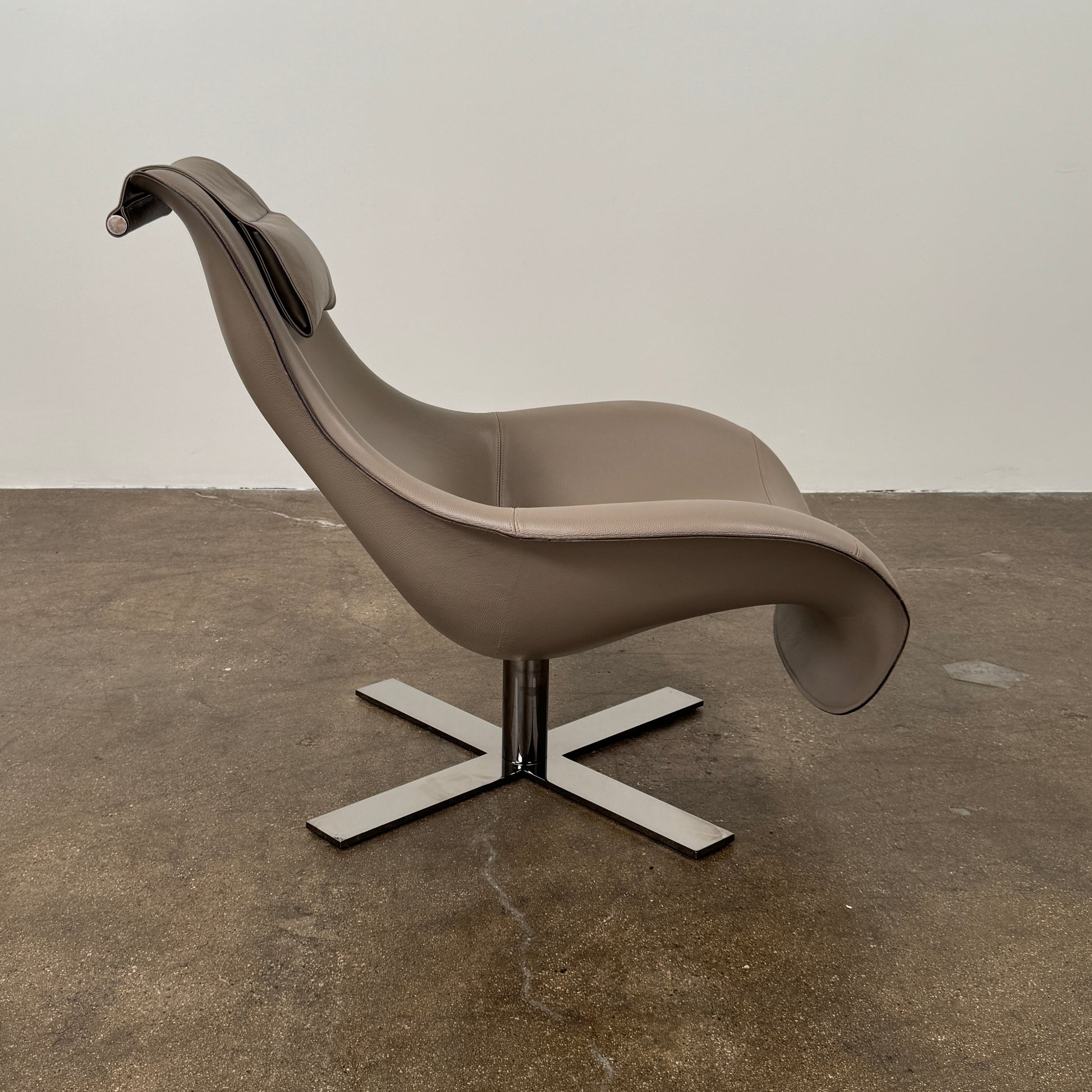 The B&B Italia Mart Armchair with Swivel Base is a modern beige lounge chair featuring a low backrest and headrest, set on a sleek swivel base against a plain white wall and concrete floor.