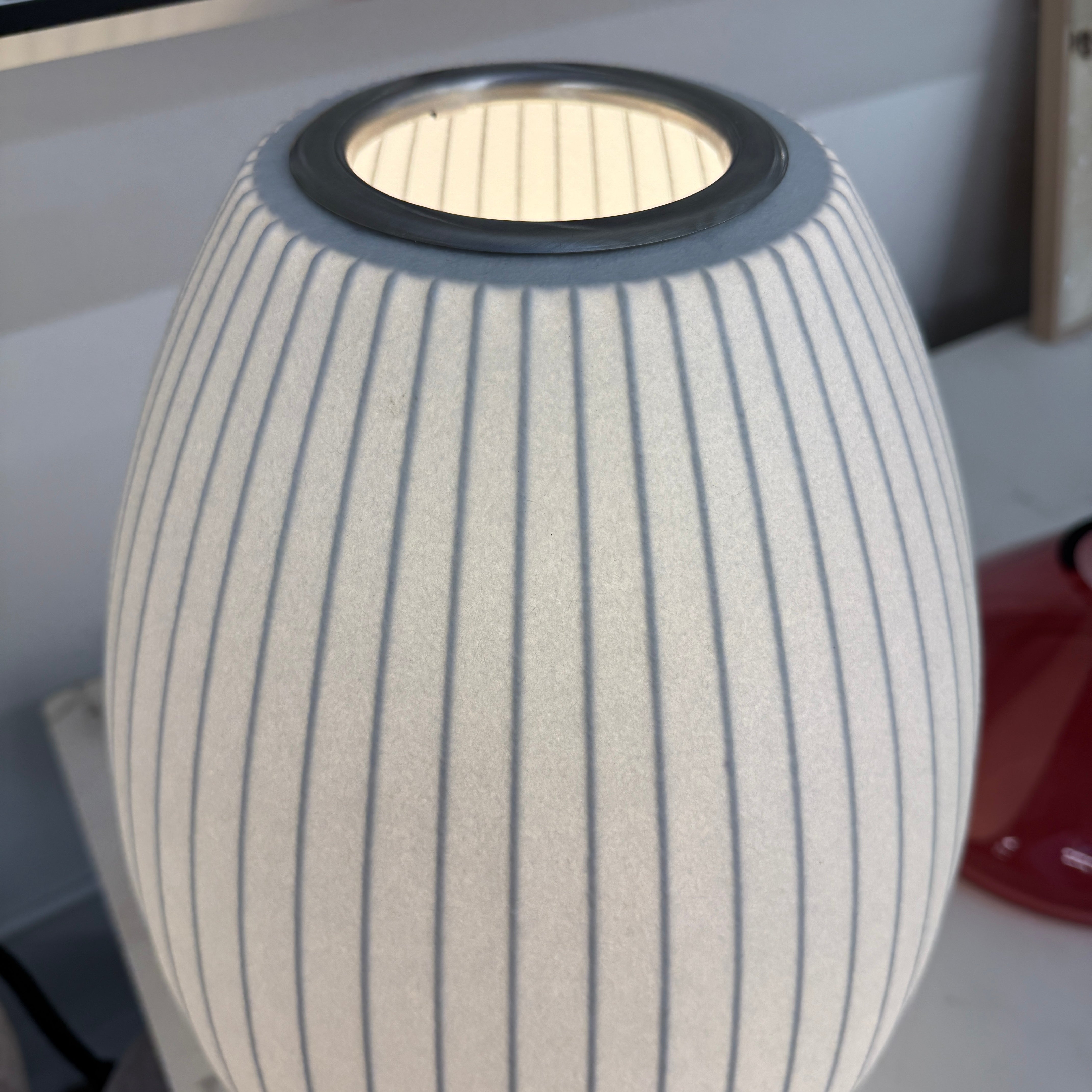 A white, oval-shaped Herman Miller Nelson Cigar Tripod Lamp with vertical ridges rests on a black surface, its cord trailing behind, while an abstract painting hangs in the background.