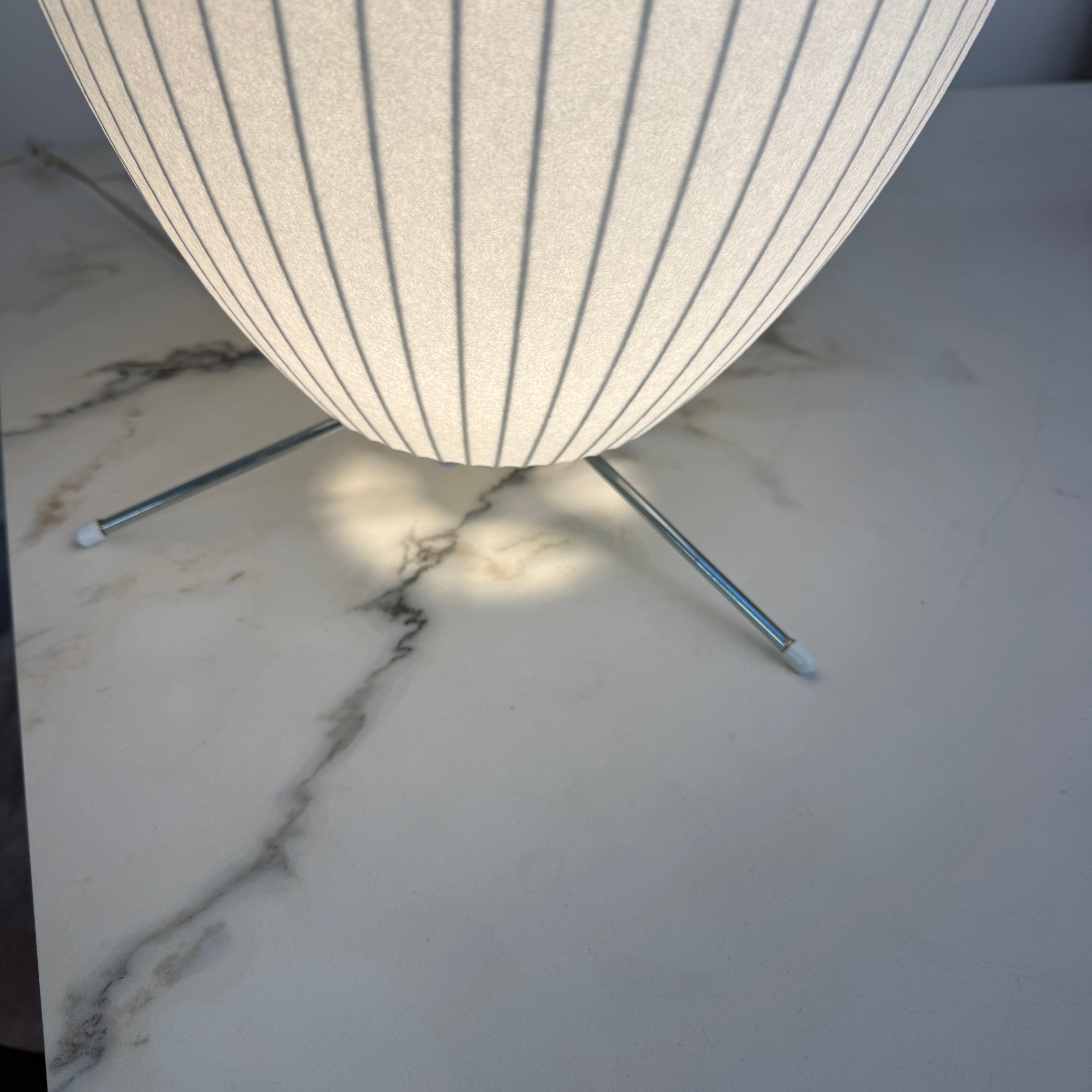 A white, oval-shaped Herman Miller Nelson Cigar Tripod Lamp with vertical ridges rests on a black surface, its cord trailing behind, while an abstract painting hangs in the background.