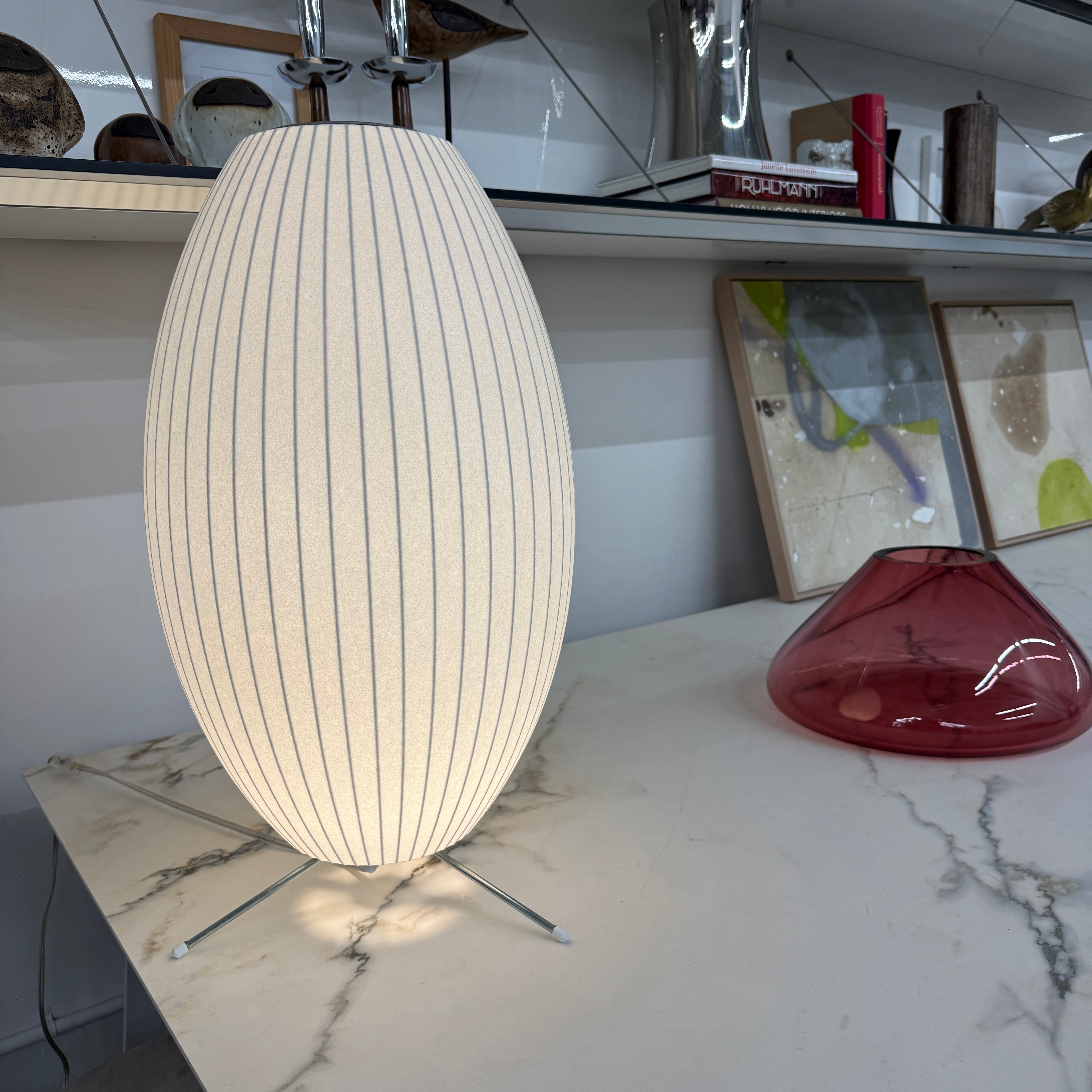 A white, oval-shaped Herman Miller Nelson Cigar Tripod Lamp with vertical ridges rests on a black surface, its cord trailing behind, while an abstract painting hangs in the background.