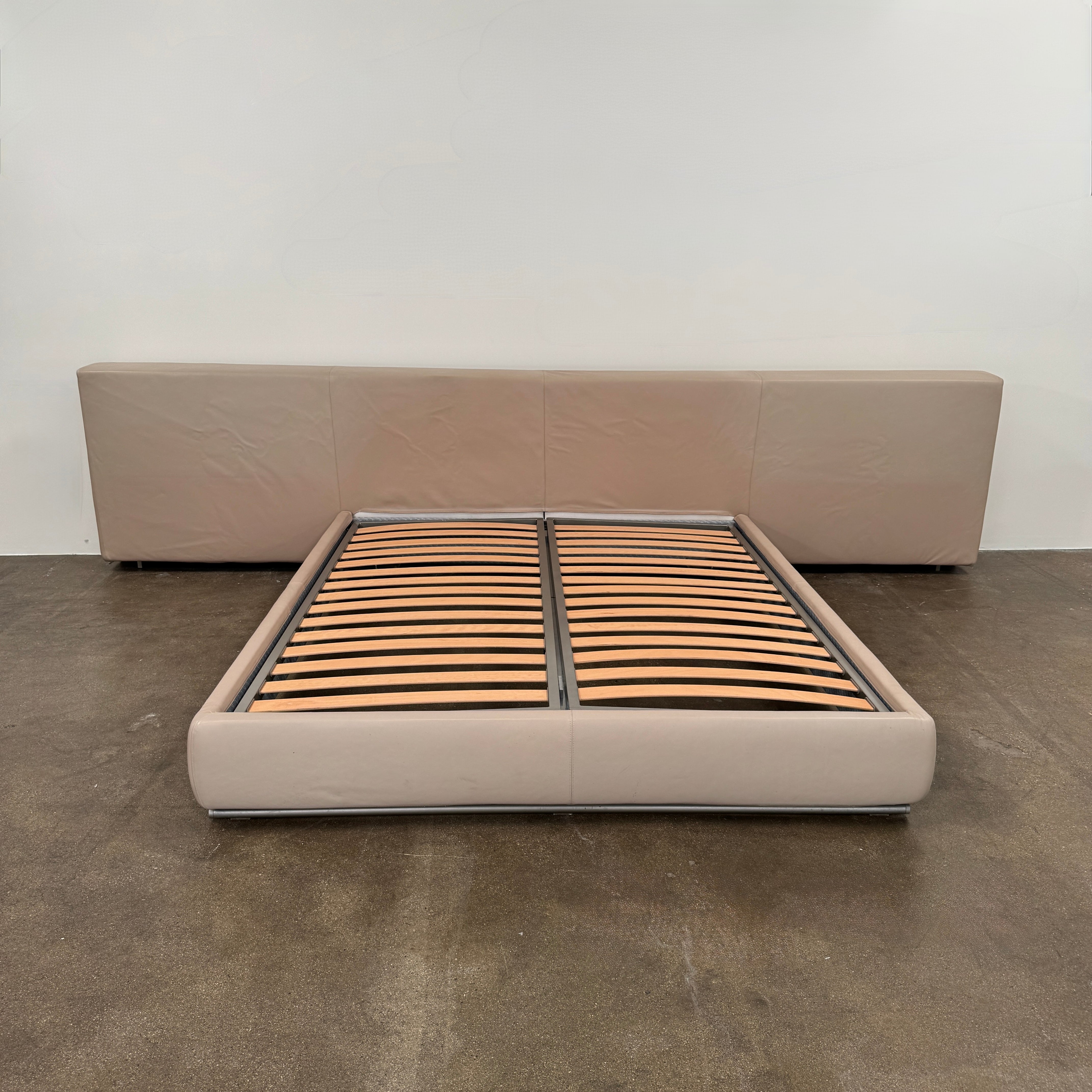 The Flexform Groundpiece Cal King Size Bed by Flexform features a beige upholstered leather frame, wide headboard, and exposed wooden slats, shown against a plain white background.