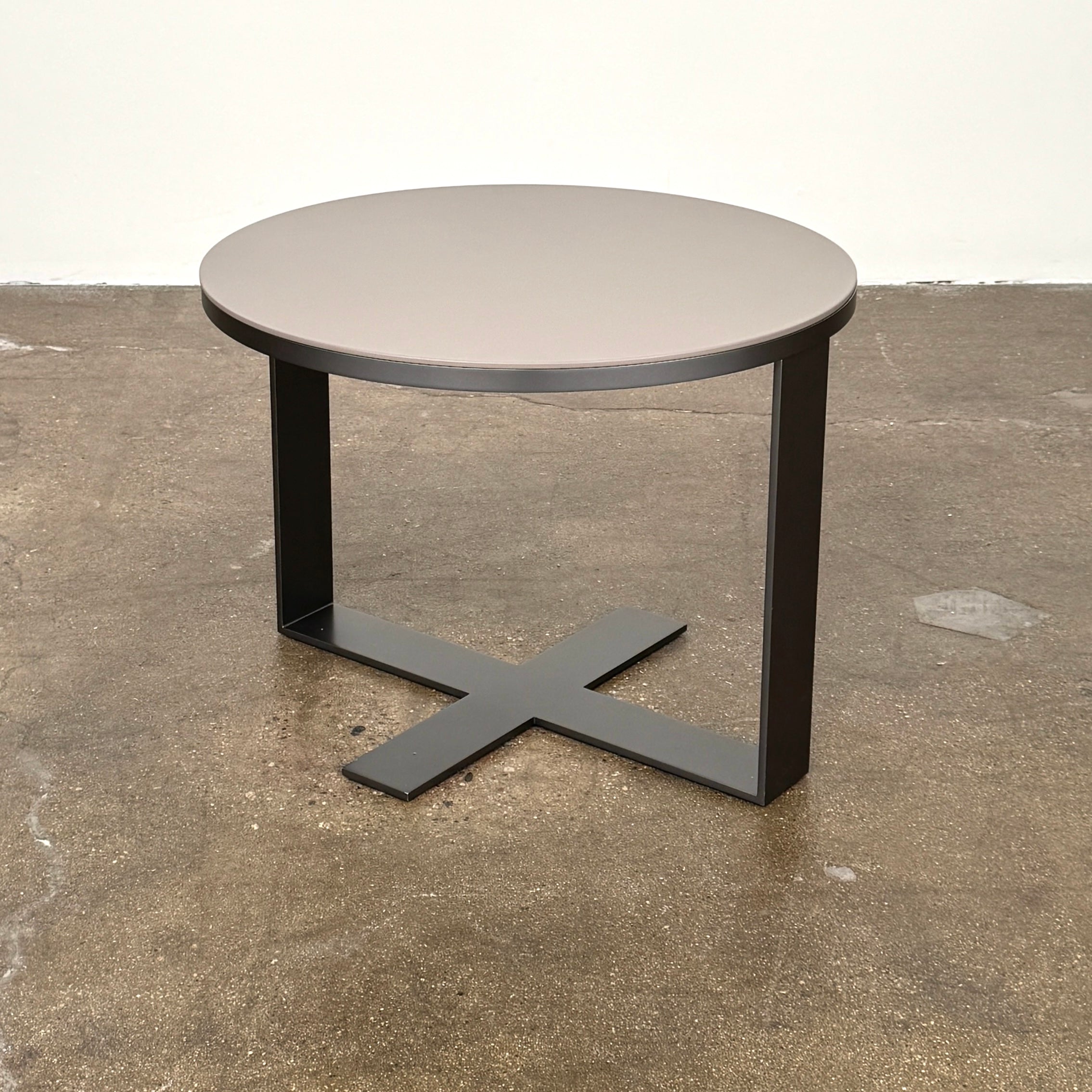 The B&B Italia Eileen Side Table, with its round gray top and single central metal leg, stands on a brown concrete floor against a plain white wall—ideal as a modern nightstand.