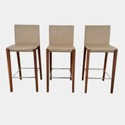 A set of three Walter Knoll Andoo Counter Stools with beige leather seats, wooden legs, and metal footrests is placed on a polished concrete floor against a plain white wall.