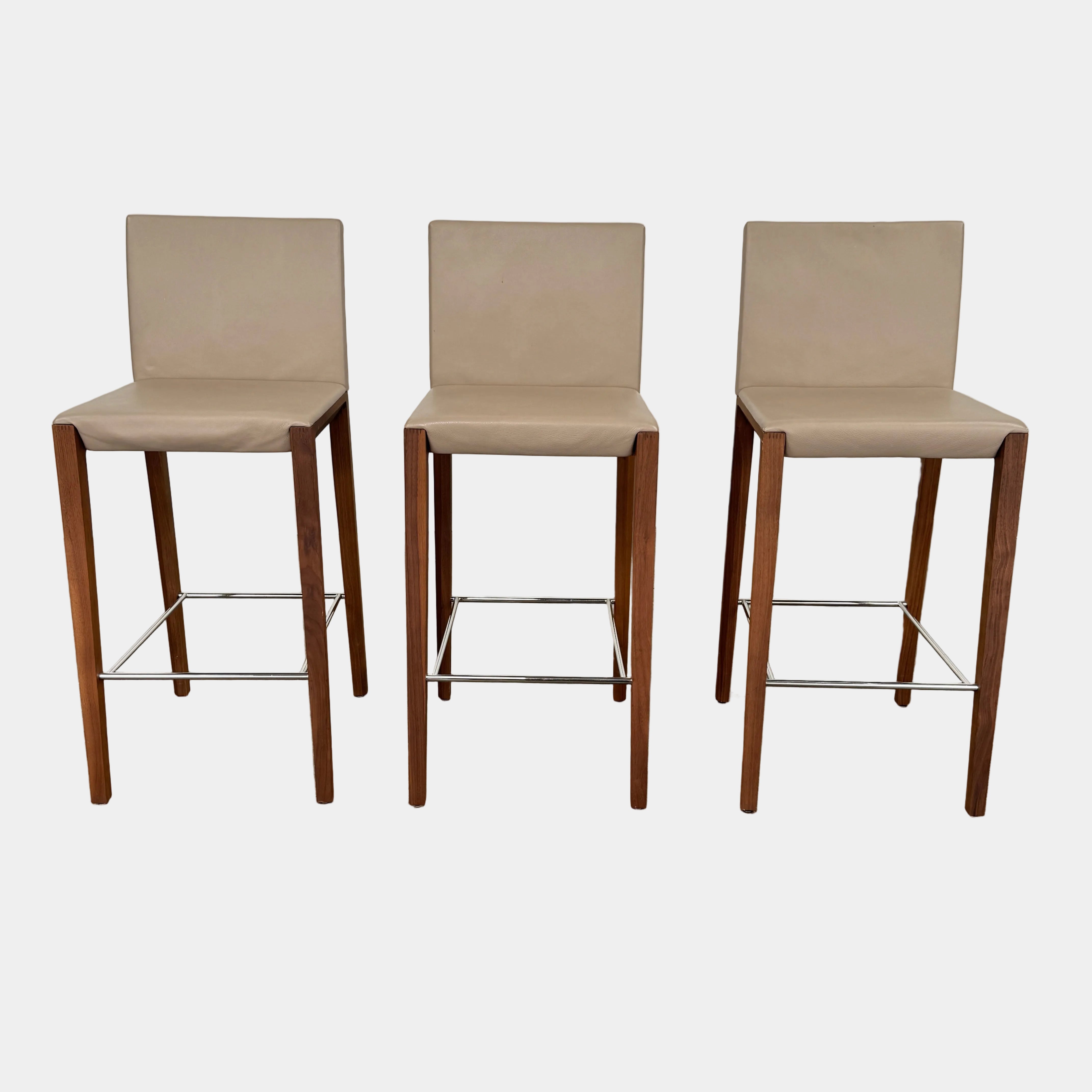 A set of three Walter Knoll Andoo Counter Stools with beige leather seats, wooden legs, and metal footrests is placed on a polished concrete floor against a plain white wall.