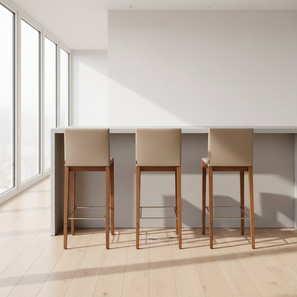 A set of three Walter Knoll Andoo Counter Stools with beige leather seats, wooden legs, and metal footrests is placed on a polished concrete floor against a plain white wall.