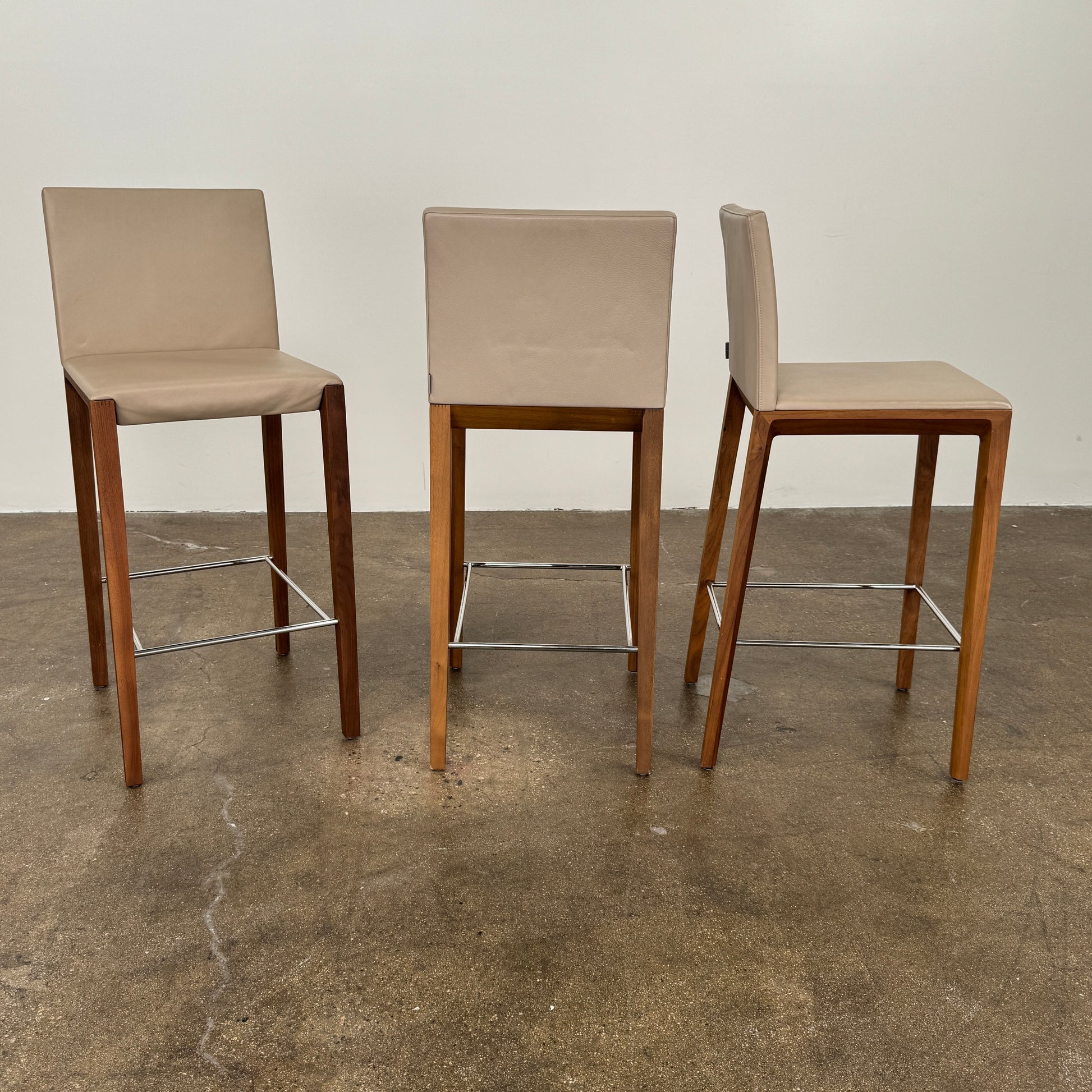 A set of three Walter Knoll Andoo Counter Stools with beige leather seats, wooden legs, and metal footrests is placed on a polished concrete floor against a plain white wall.