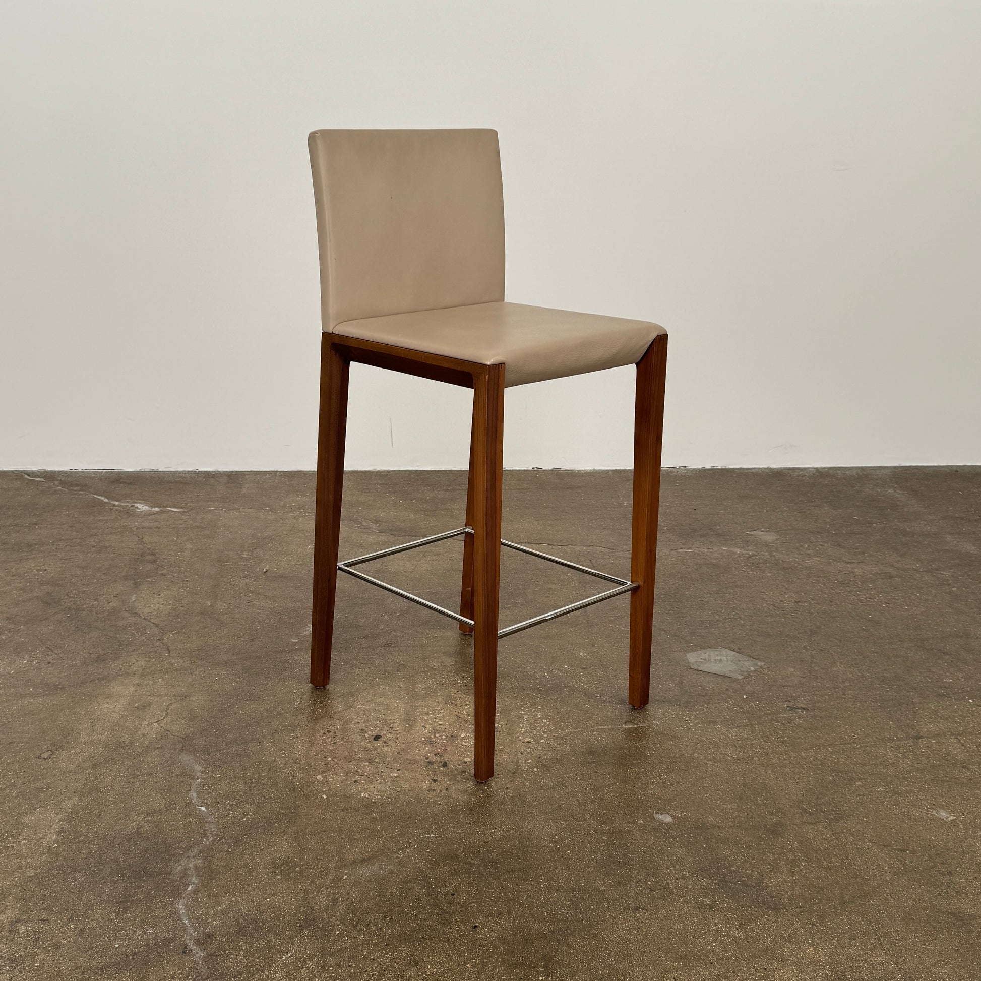 A set of three Walter Knoll Andoo Counter Stools with beige leather seats, wooden legs, and metal footrests is placed on a polished concrete floor against a plain white wall.