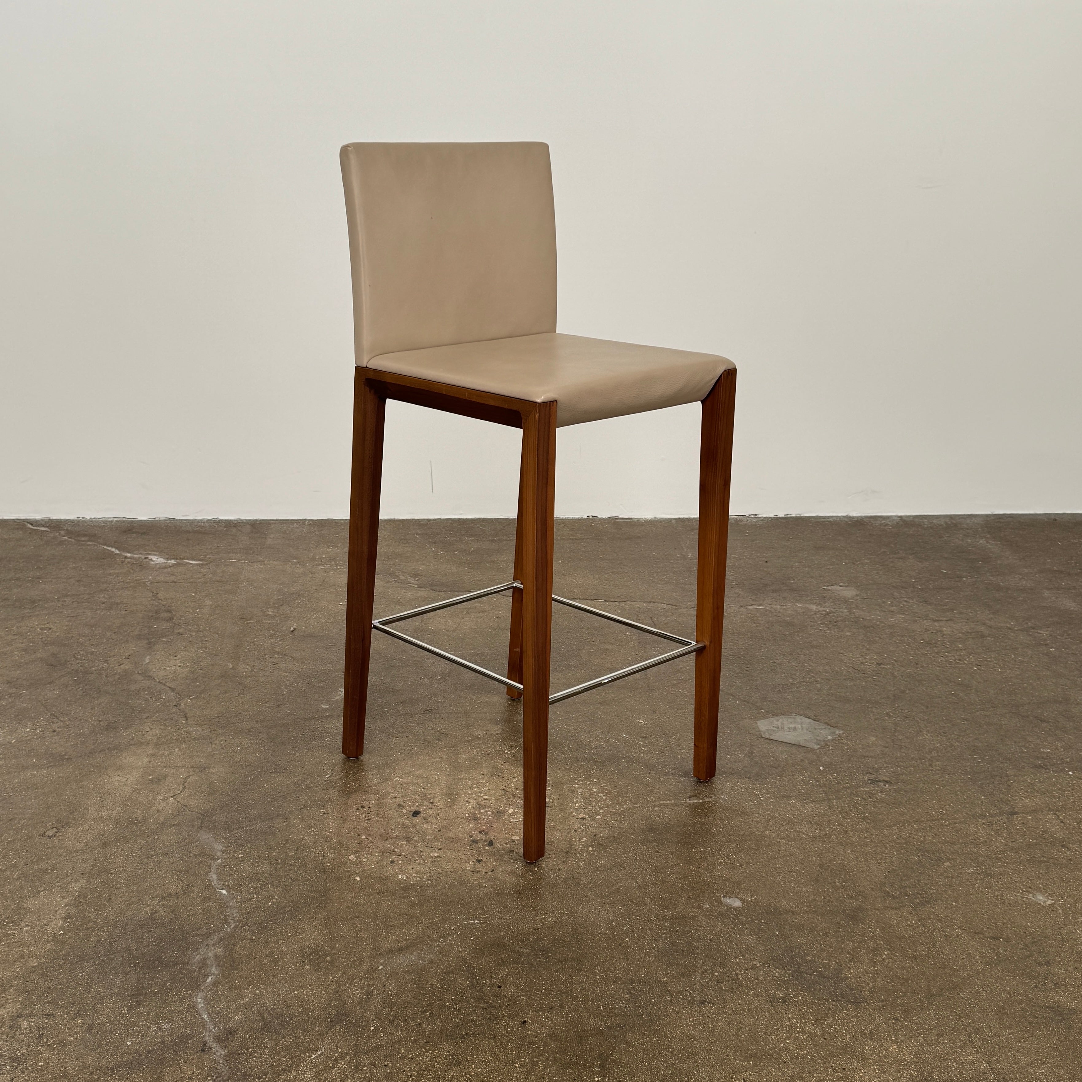 A set of three Walter Knoll Andoo Counter Stools with beige leather seats, wooden legs, and metal footrests is placed on a polished concrete floor against a plain white wall.