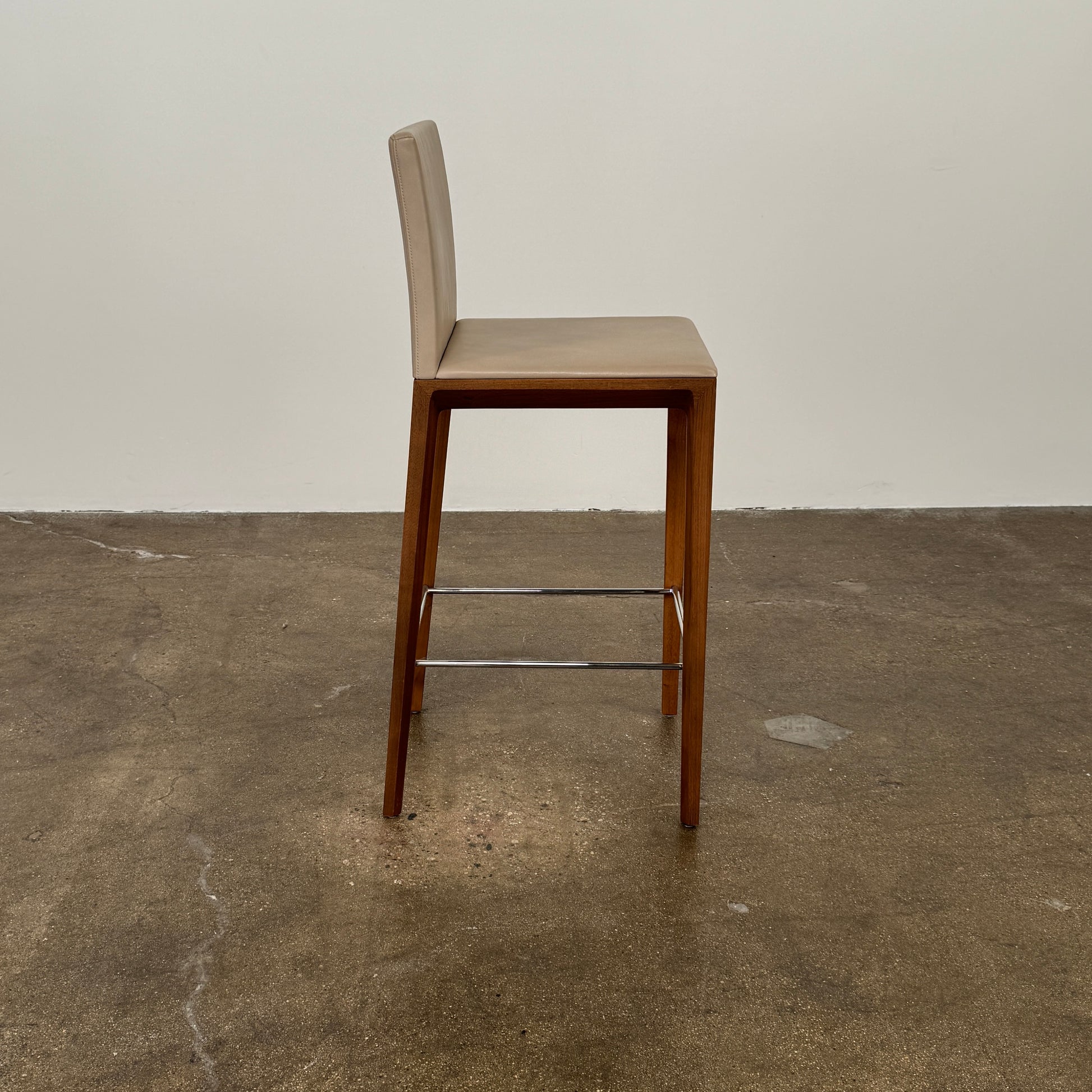 A set of three Walter Knoll Andoo Counter Stools with beige leather seats, wooden legs, and metal footrests is placed on a polished concrete floor against a plain white wall.