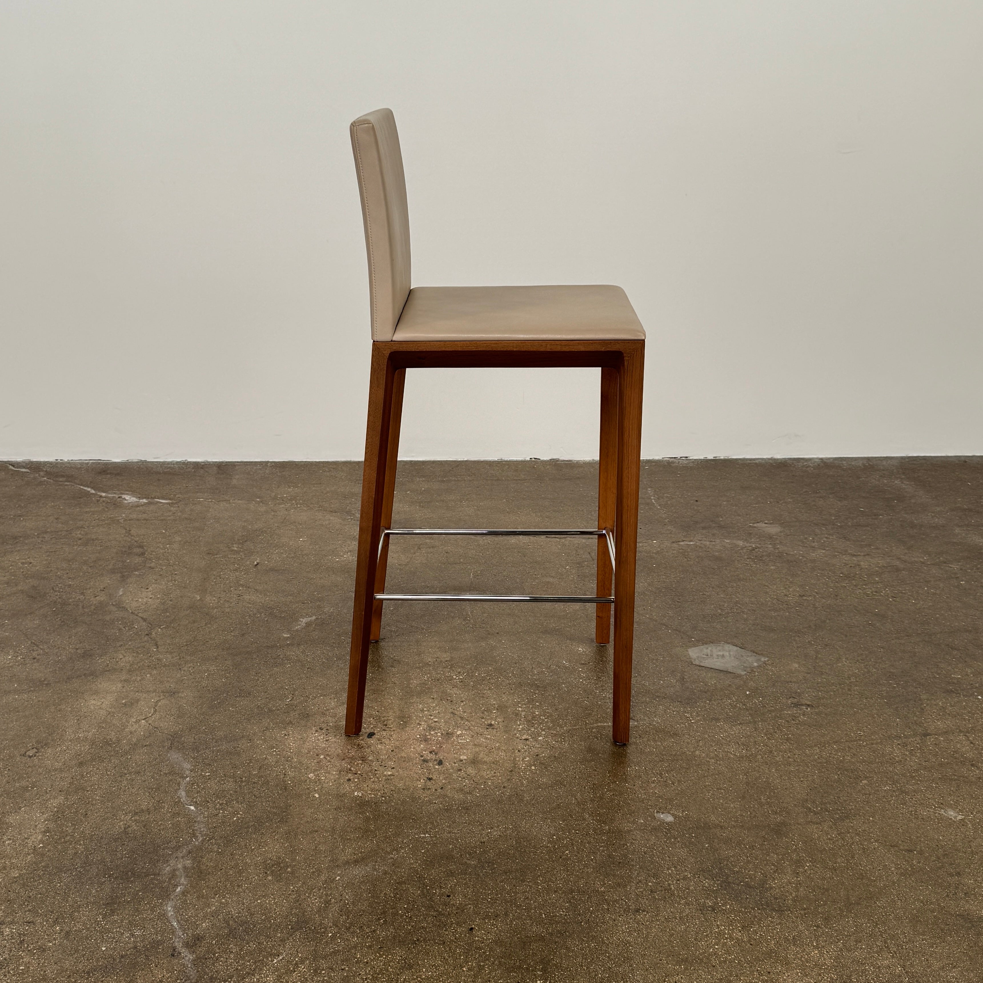A set of three Walter Knoll Andoo Counter Stools with beige leather seats, wooden legs, and metal footrests is placed on a polished concrete floor against a plain white wall.