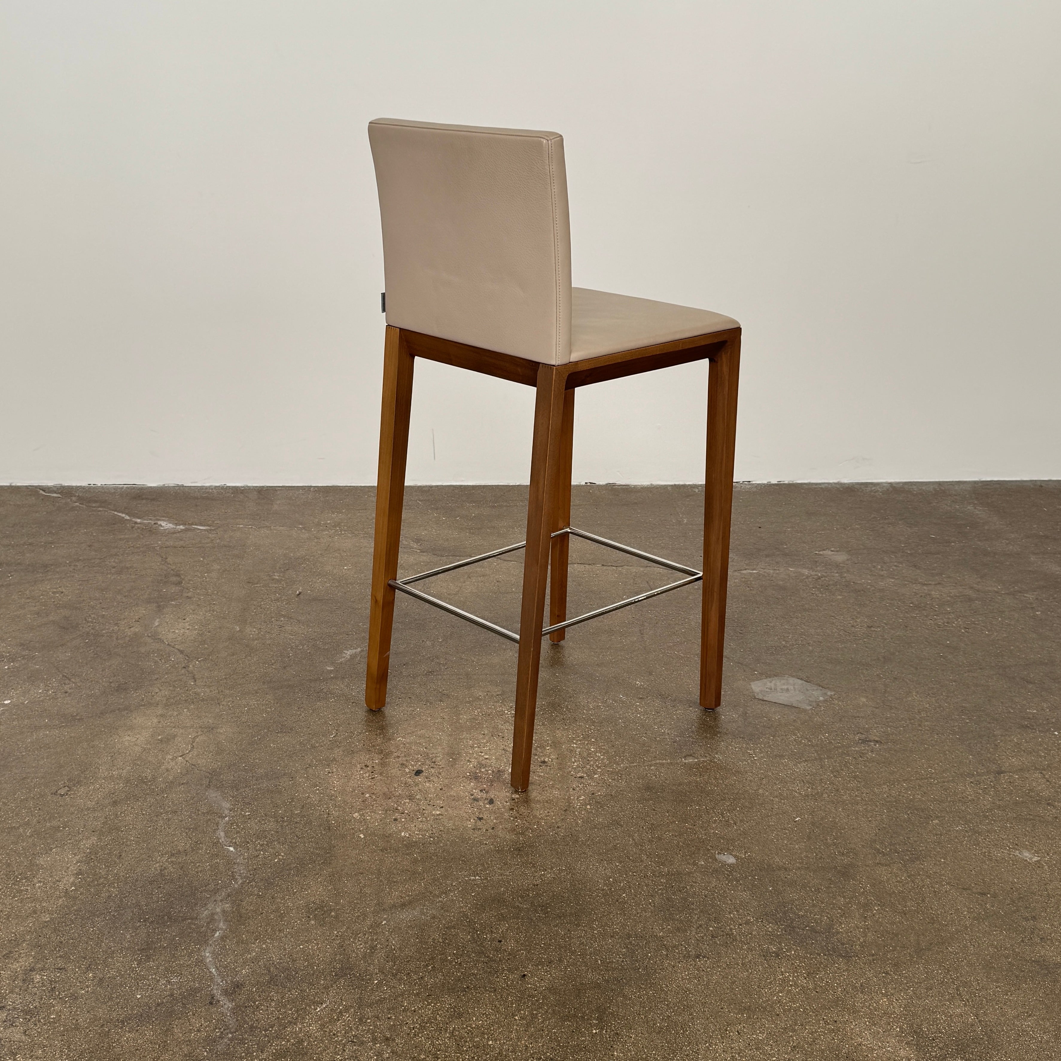 A set of three Walter Knoll Andoo Counter Stools with beige leather seats, wooden legs, and metal footrests is placed on a polished concrete floor against a plain white wall.