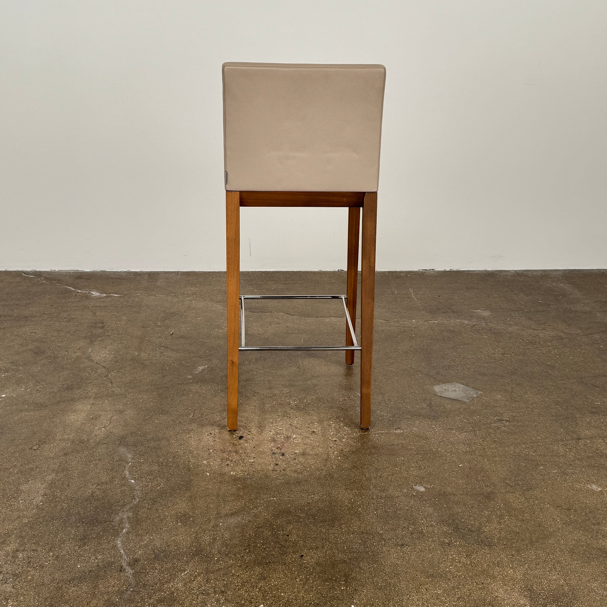 A set of three Walter Knoll Andoo Counter Stools with beige leather seats, wooden legs, and metal footrests is placed on a polished concrete floor against a plain white wall.