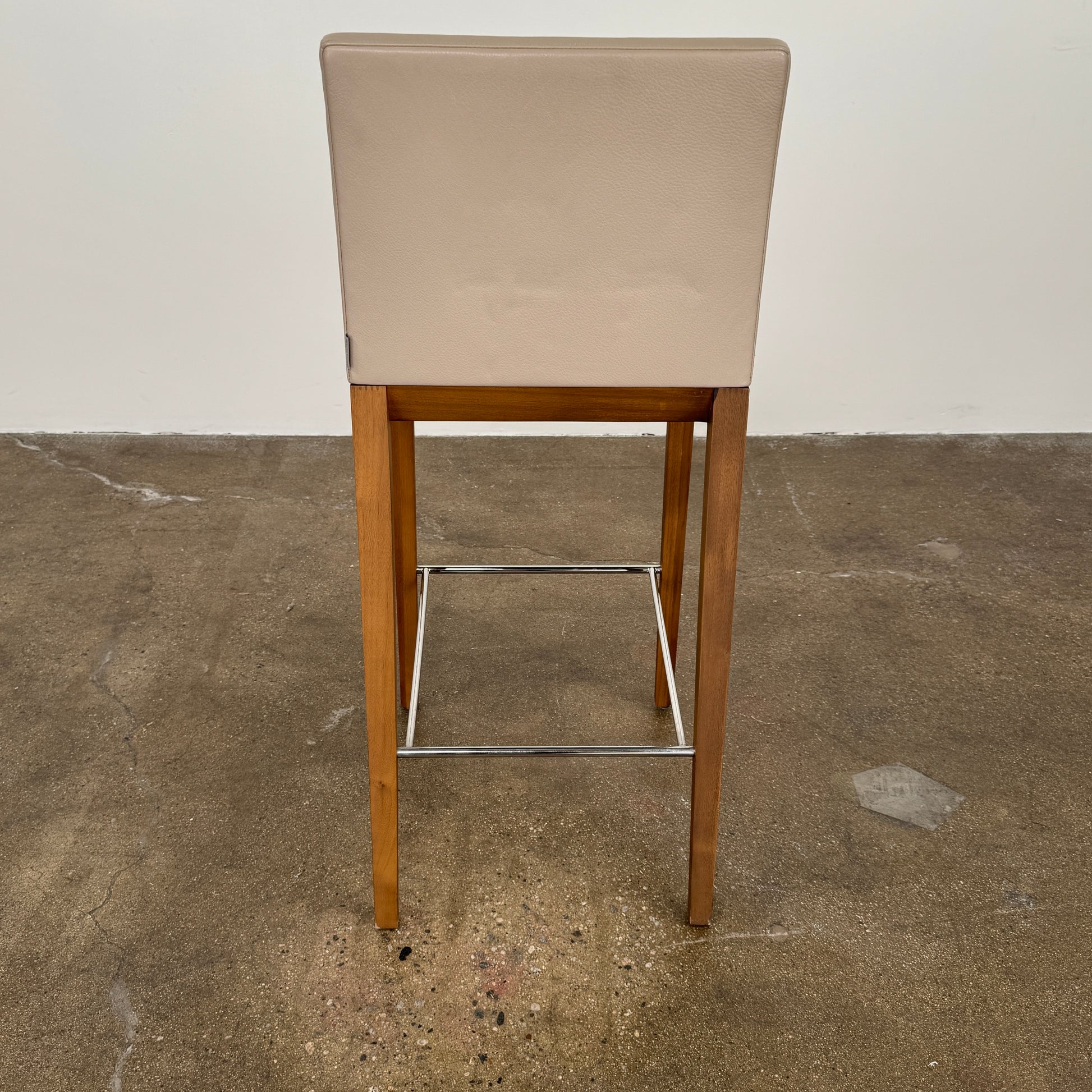 A set of three Walter Knoll Andoo Counter Stools with beige leather seats, wooden legs, and metal footrests is placed on a polished concrete floor against a plain white wall.
