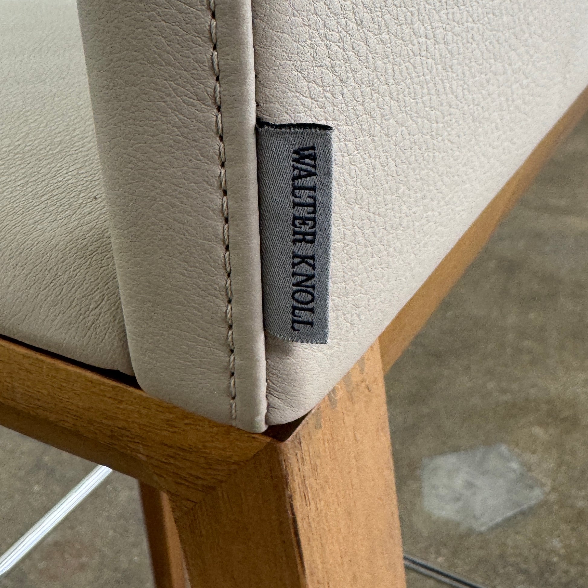 A set of three Walter Knoll Andoo Counter Stools with beige leather seats, wooden legs, and metal footrests is placed on a polished concrete floor against a plain white wall.