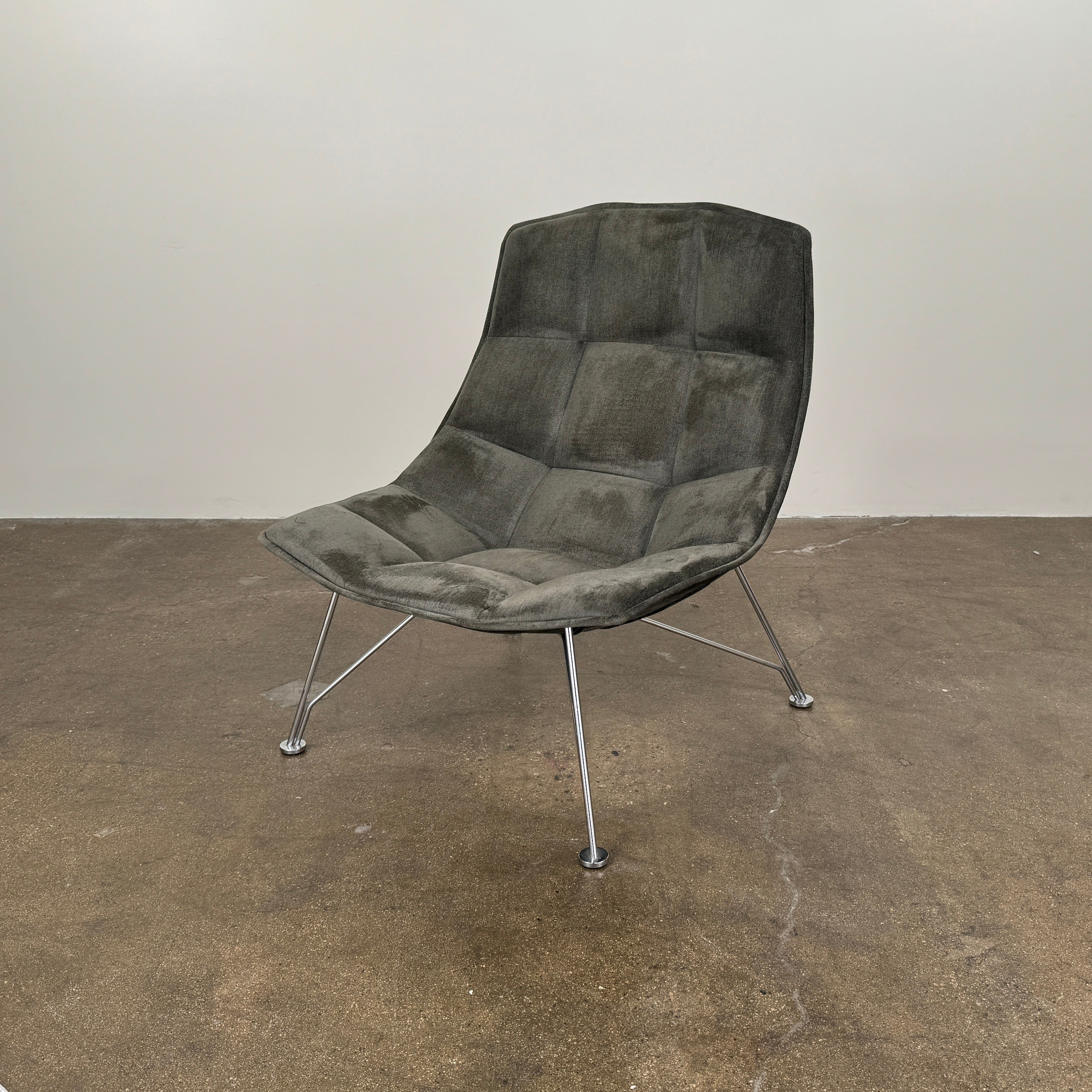 The Knoll Jehs and Laub Lounge Chair by Knoll, featuring a dark tufted fabric seat and chrome legs, is placed on a polished concrete floor against a plain white wall.