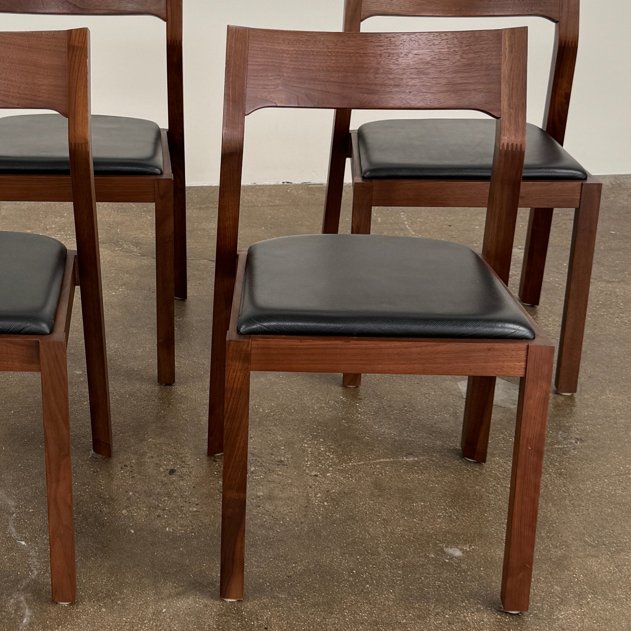 The Case Profile Dining Chair Set of 4 by Case, featuring wooden frames and black cushioned seats, is arranged on a concrete floor against a plain off-white wall.