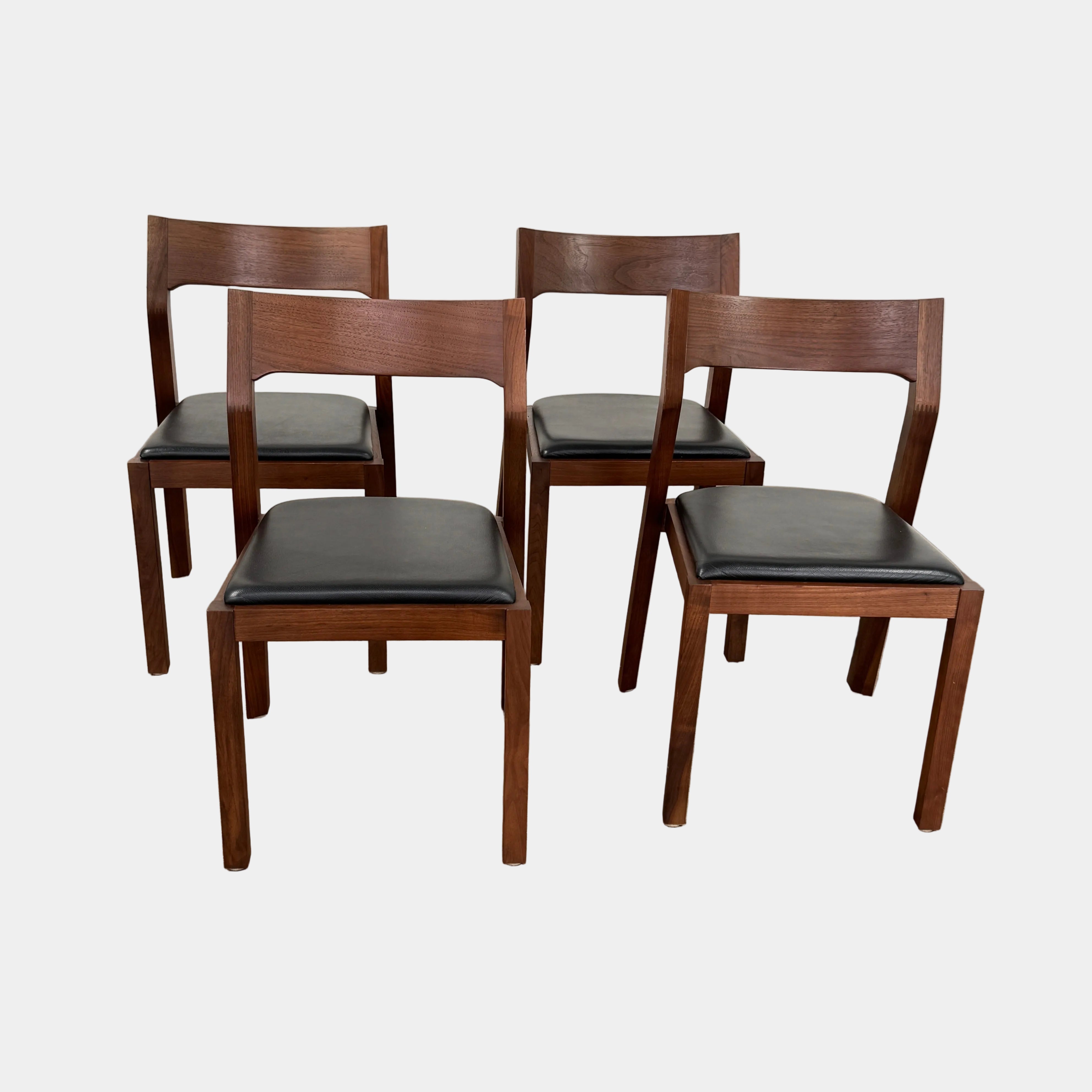 The Case Profile Dining Chair Set of 4 by Case, featuring wooden frames and black cushioned seats, is arranged on a concrete floor against a plain off-white wall.