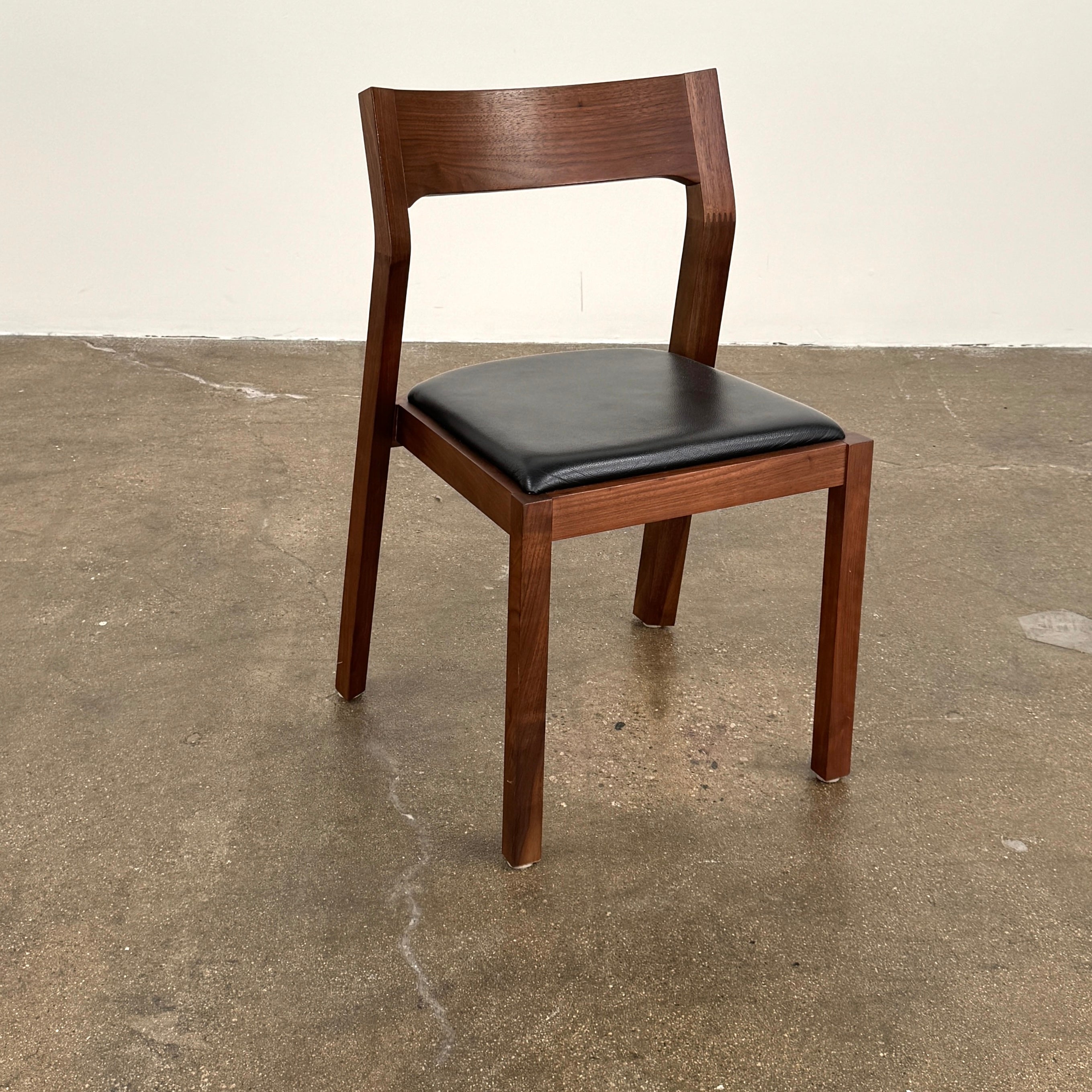 The Case Profile Dining Chair Set of 4 by Case, featuring wooden frames and black cushioned seats, is arranged on a concrete floor against a plain off-white wall.