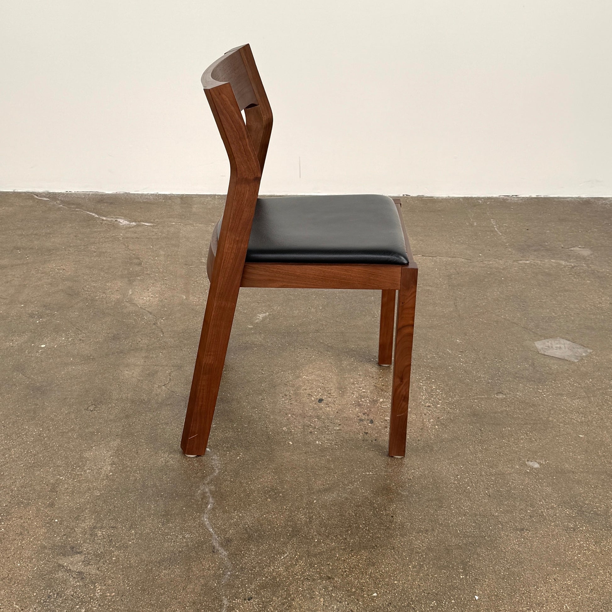 The Case Profile Dining Chair Set of 4 by Case, featuring wooden frames and black cushioned seats, is arranged on a concrete floor against a plain off-white wall.
