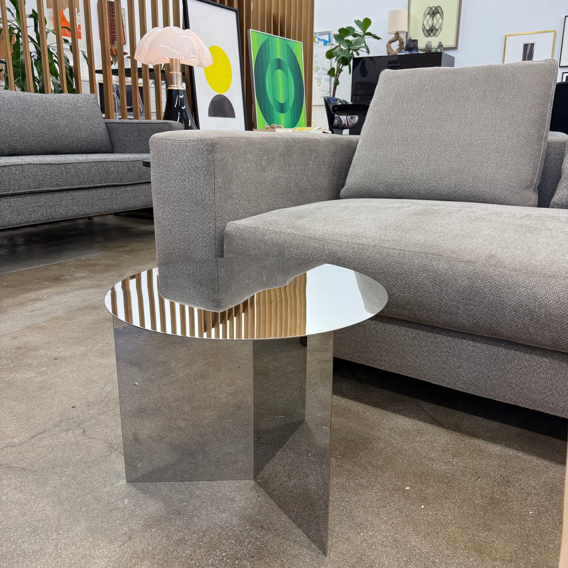 The Hay Slit Side Table by Hay features a round, reflective design with mirrored sides and is shown atop a dark wooden surface against a plain white wall.