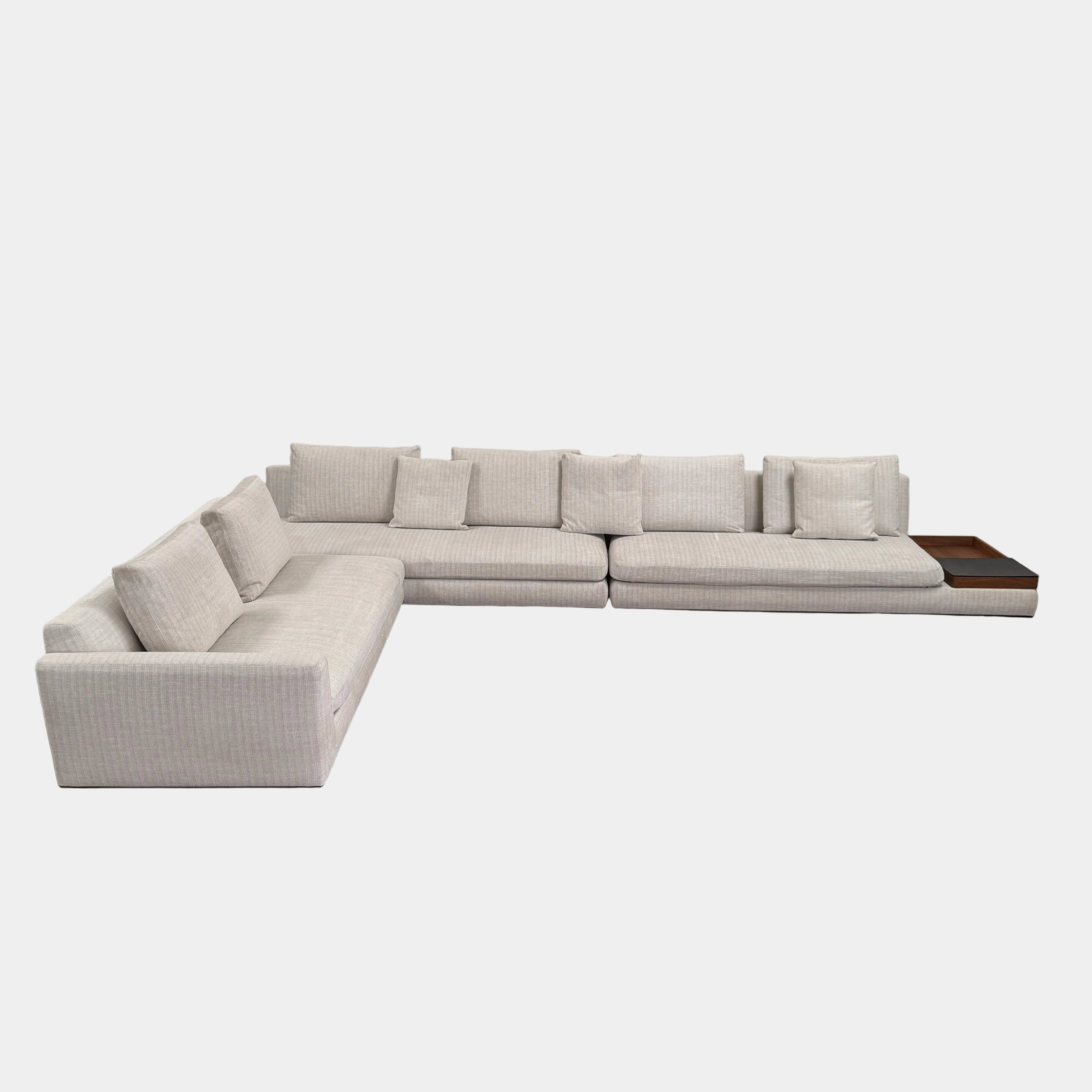 The Walter Knoll Tama Living Modular Sofa, upholstered in light gray luxury fabric with a chaise lounge and an integrated wooden side table, stands on a bare concrete floor against a plain white wall.