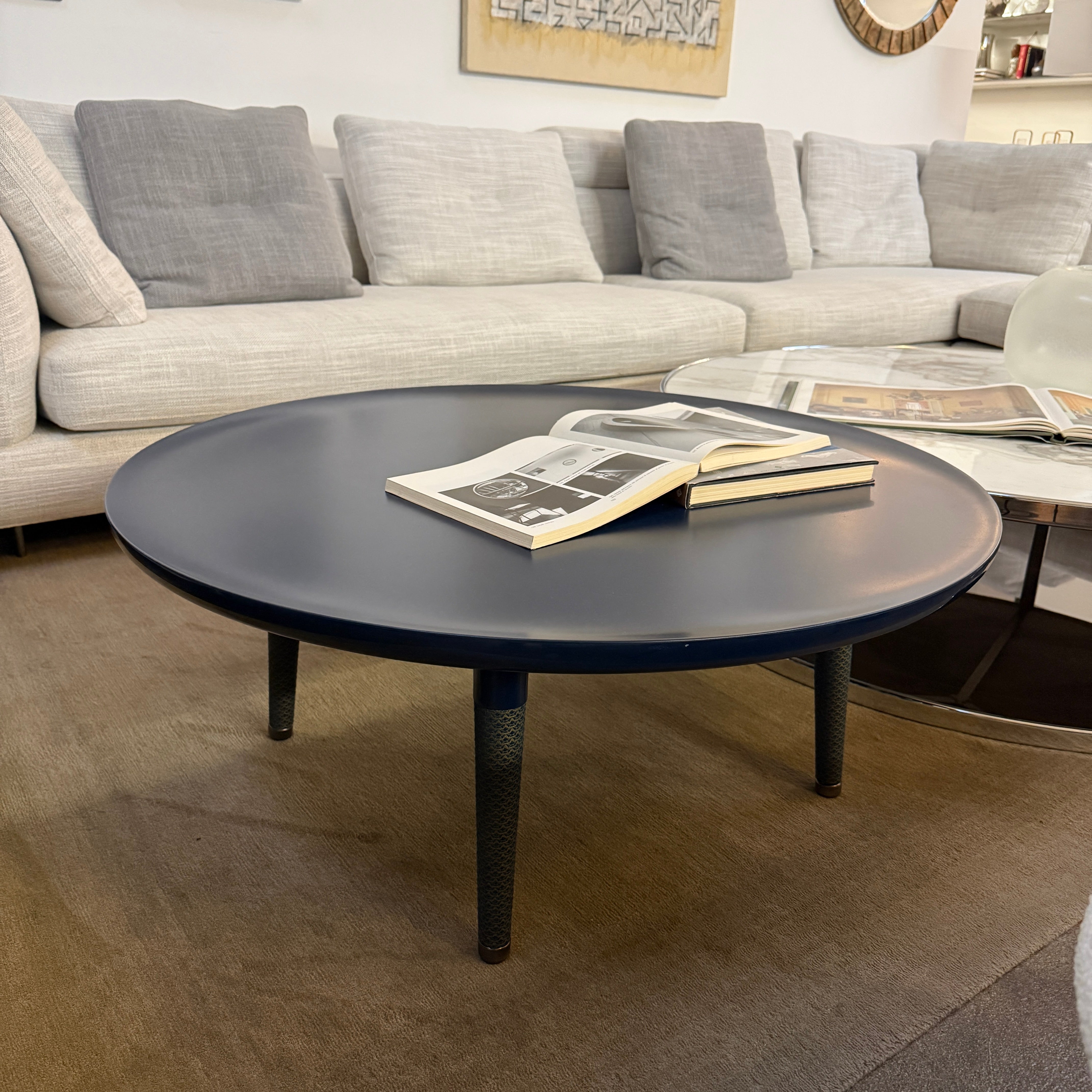 The Roche Bobois Globe Trotter La Parisienne cocktail table features a round, dark blue top and three textured legs, shown against a light gray background.