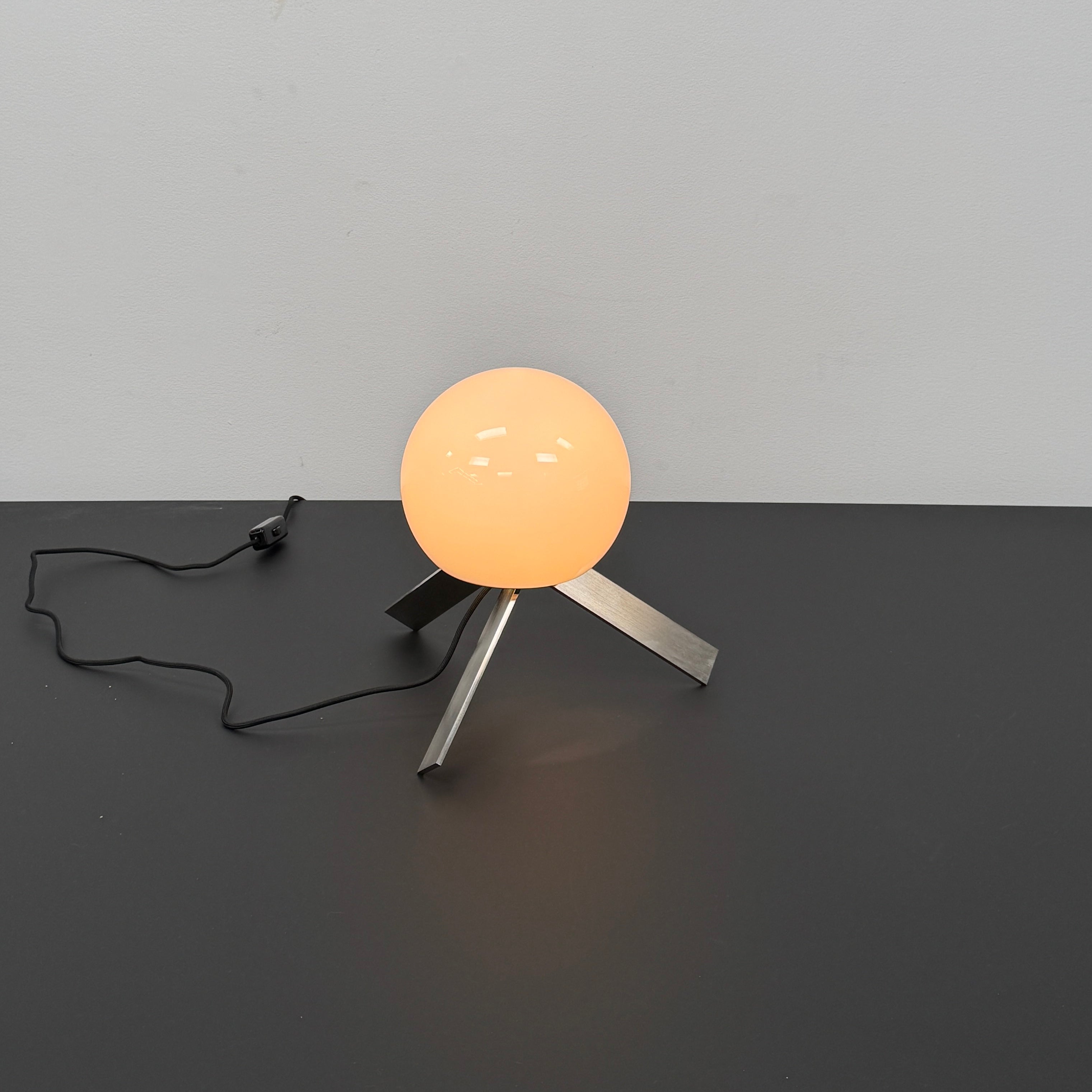 The Michael Anastassiades To The Top Table Lamp features a spherical white opaline glass shade and sits on three gold angled legs, designed by Michael Anastassiades, set against a plain light background.
