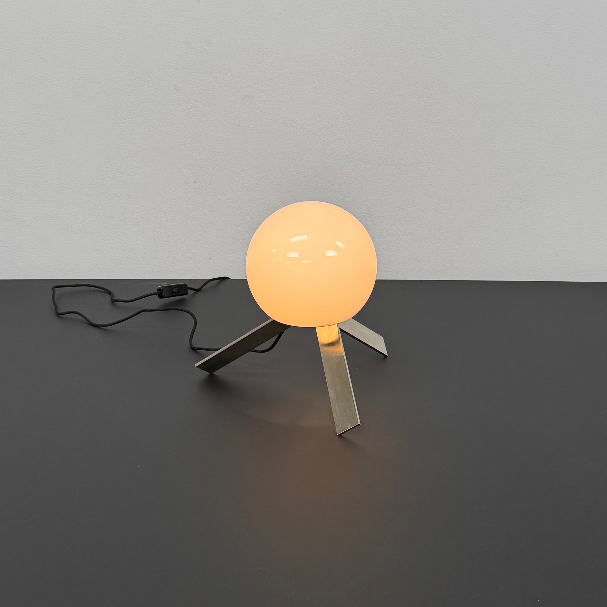 The Michael Anastassiades To The Top Table Lamp features a spherical white opaline glass shade and sits on three gold angled legs, designed by Michael Anastassiades, set against a plain light background.