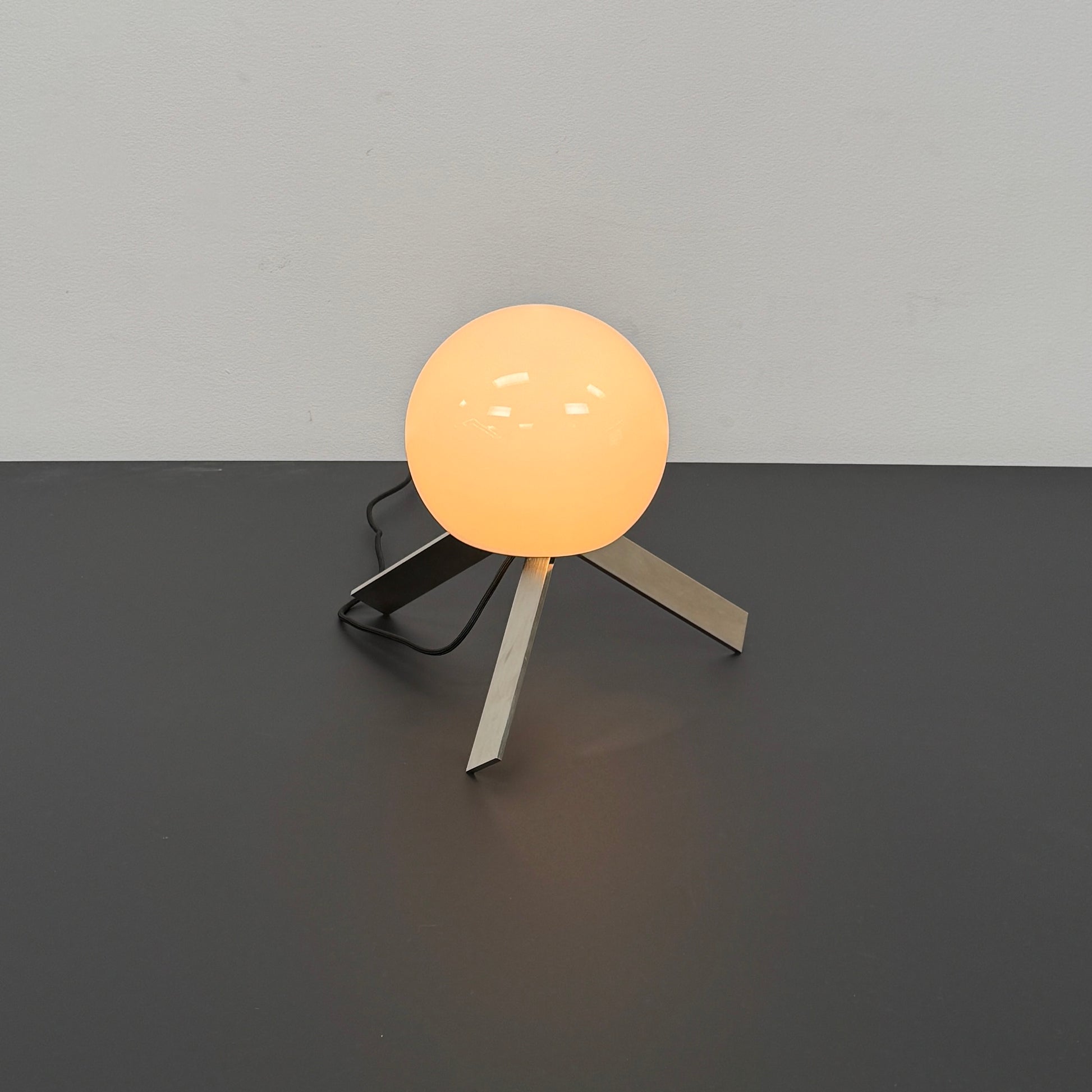 The Michael Anastassiades To The Top Table Lamp features a spherical white opaline glass shade and sits on three gold angled legs, designed by Michael Anastassiades, set against a plain light background.
