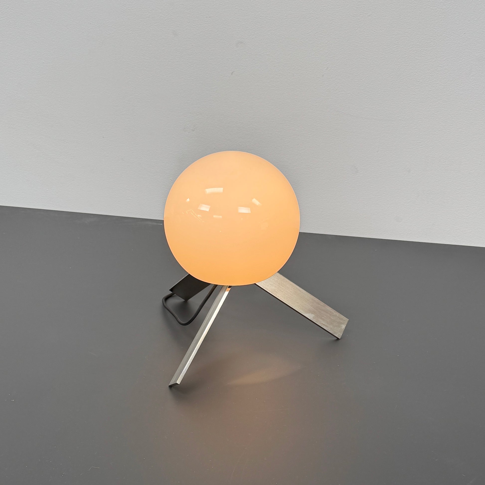 The Michael Anastassiades To The Top Table Lamp features a spherical white opaline glass shade and sits on three gold angled legs, designed by Michael Anastassiades, set against a plain light background.