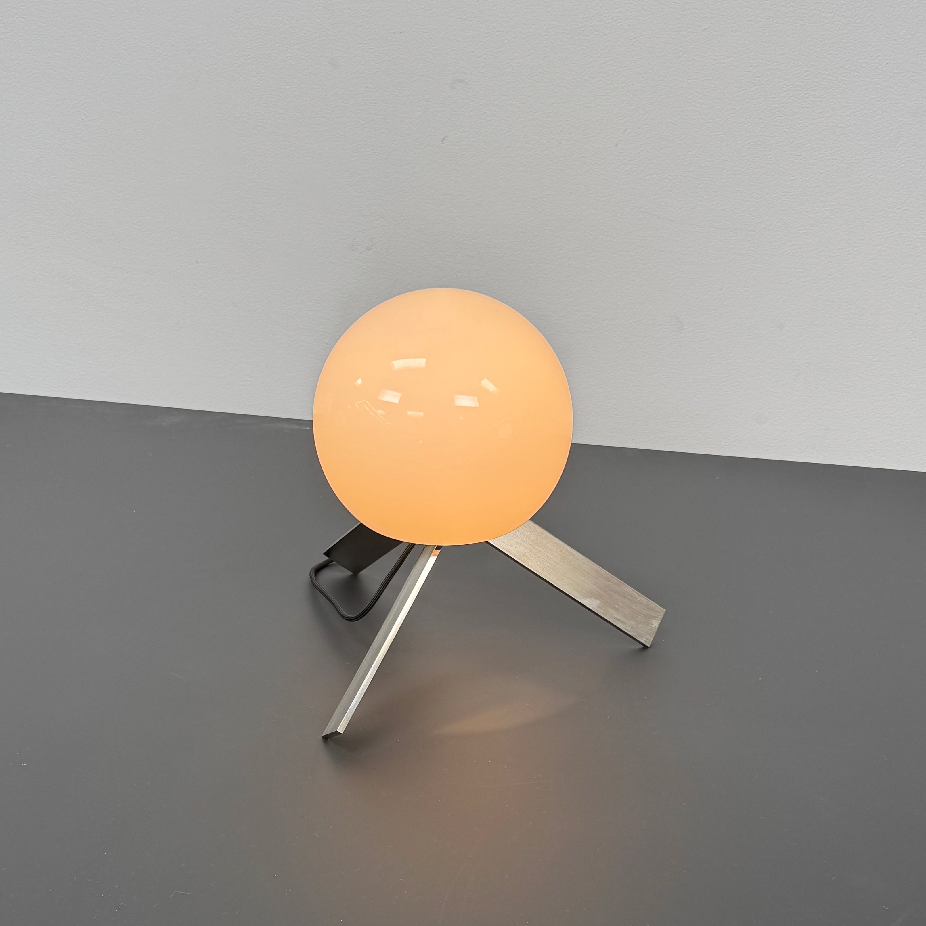 The Michael Anastassiades To The Top Table Lamp features a spherical white opaline glass shade and sits on three gold angled legs, designed by Michael Anastassiades, set against a plain light background.