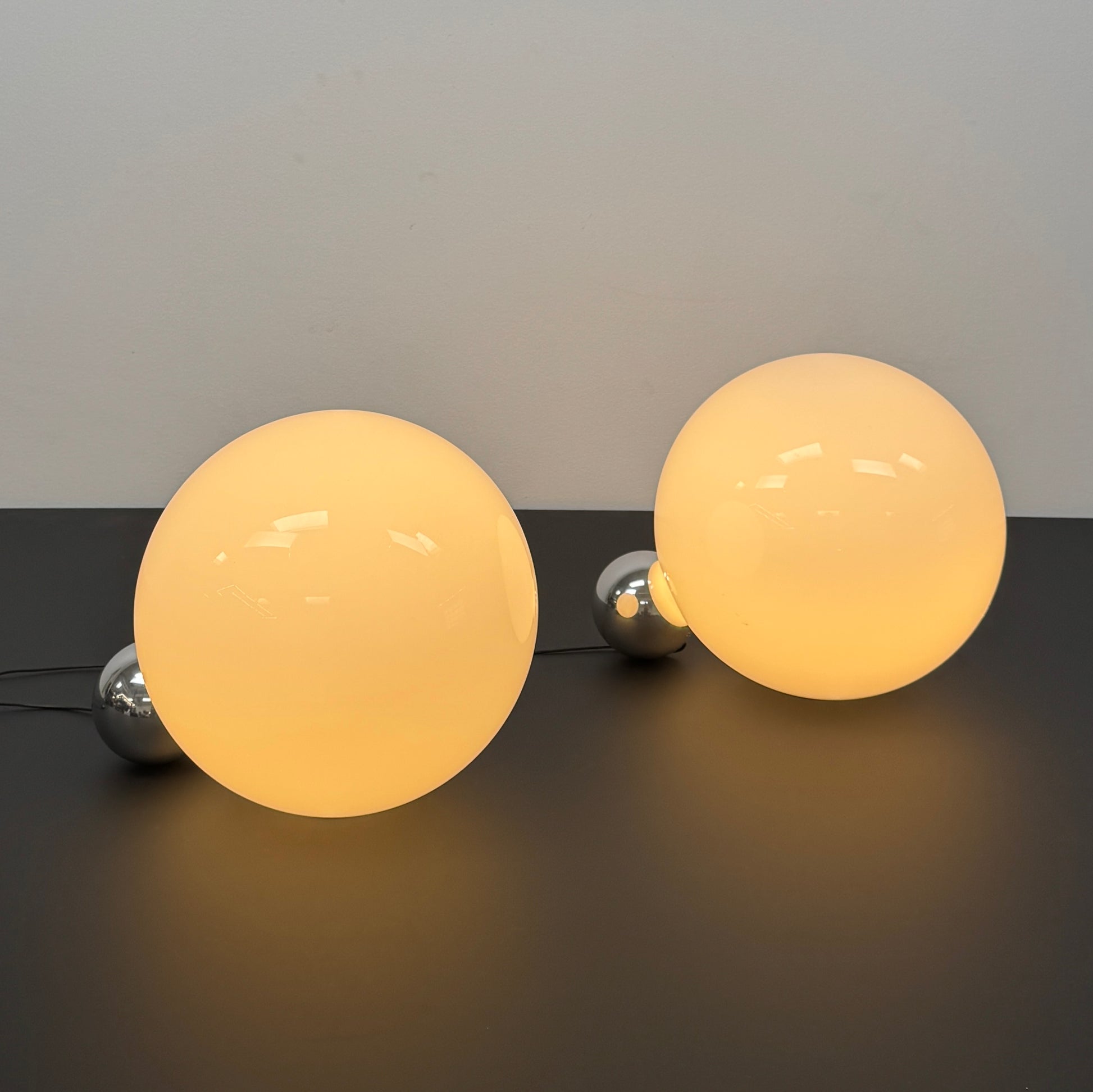 Two Flos Copycat Table Lamps by Flos, featuring glowing frosted glass spheres and chrome bases, make a sculptural statement on a dark surface against a light-colored wall.