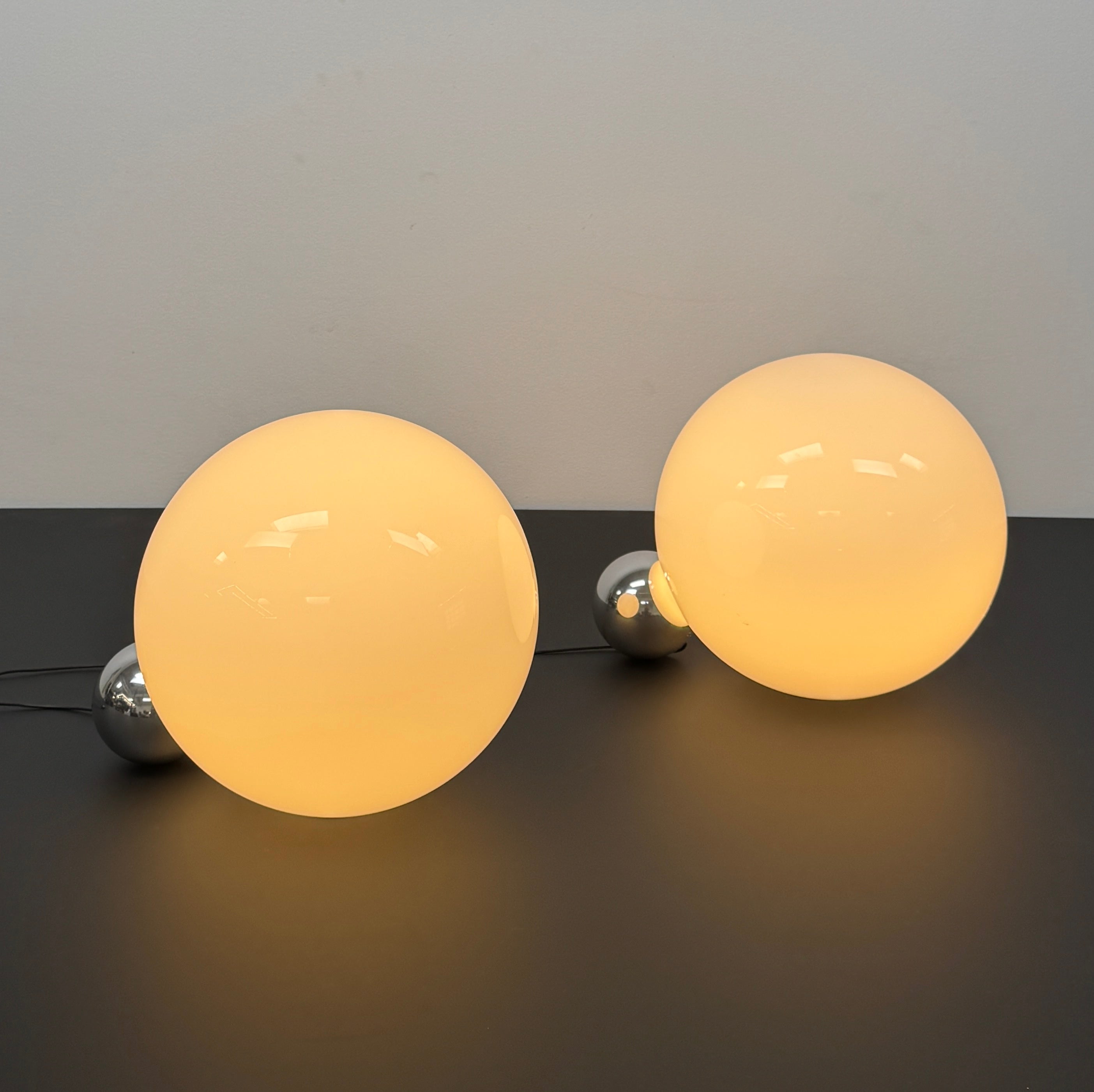 Two Flos Copycat Table Lamps by Flos, featuring glowing frosted glass spheres and chrome bases, make a sculptural statement on a dark surface against a light-colored wall.