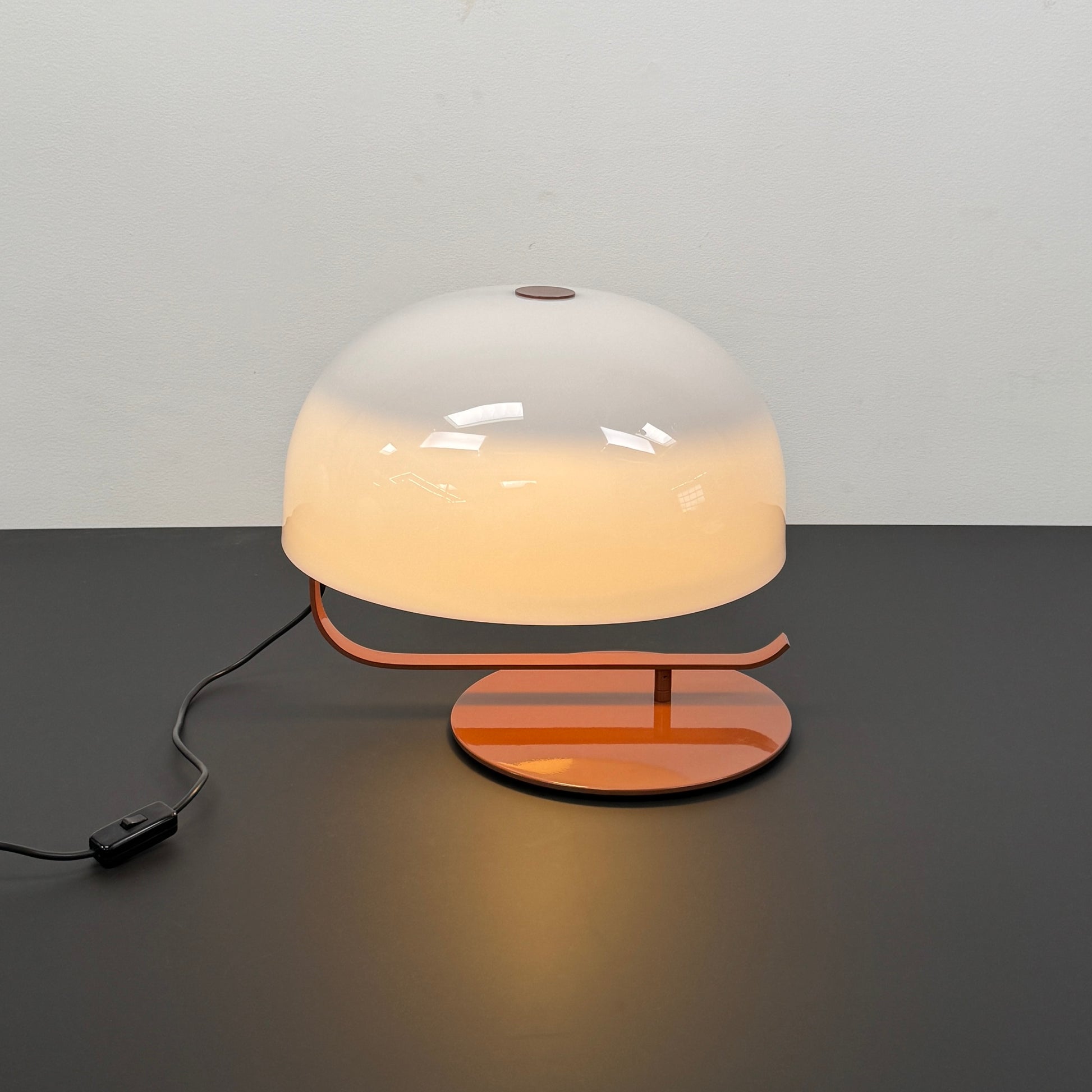 The Oluce Zanuso 275 Table Light by Oluce is a modern lamp inspired by Italian design, with a white domed glass shade, orange base, and black power cord with switch, shown against a plain background.