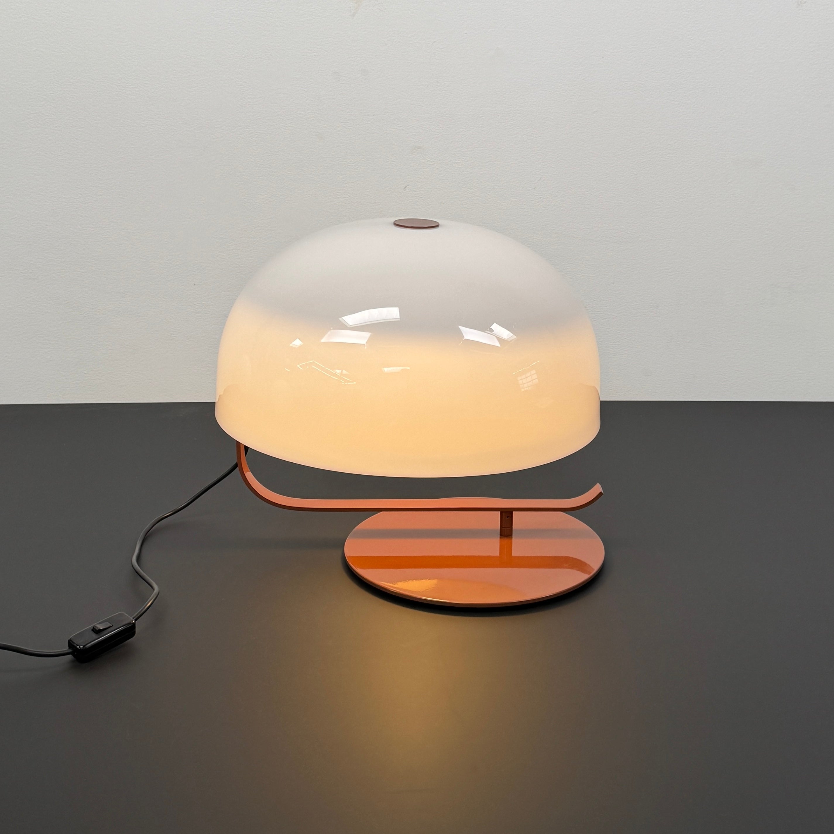 The Oluce Zanuso 275 Table Light by Oluce is a modern lamp inspired by Italian design, with a white domed glass shade, orange base, and black power cord with switch, shown against a plain background.