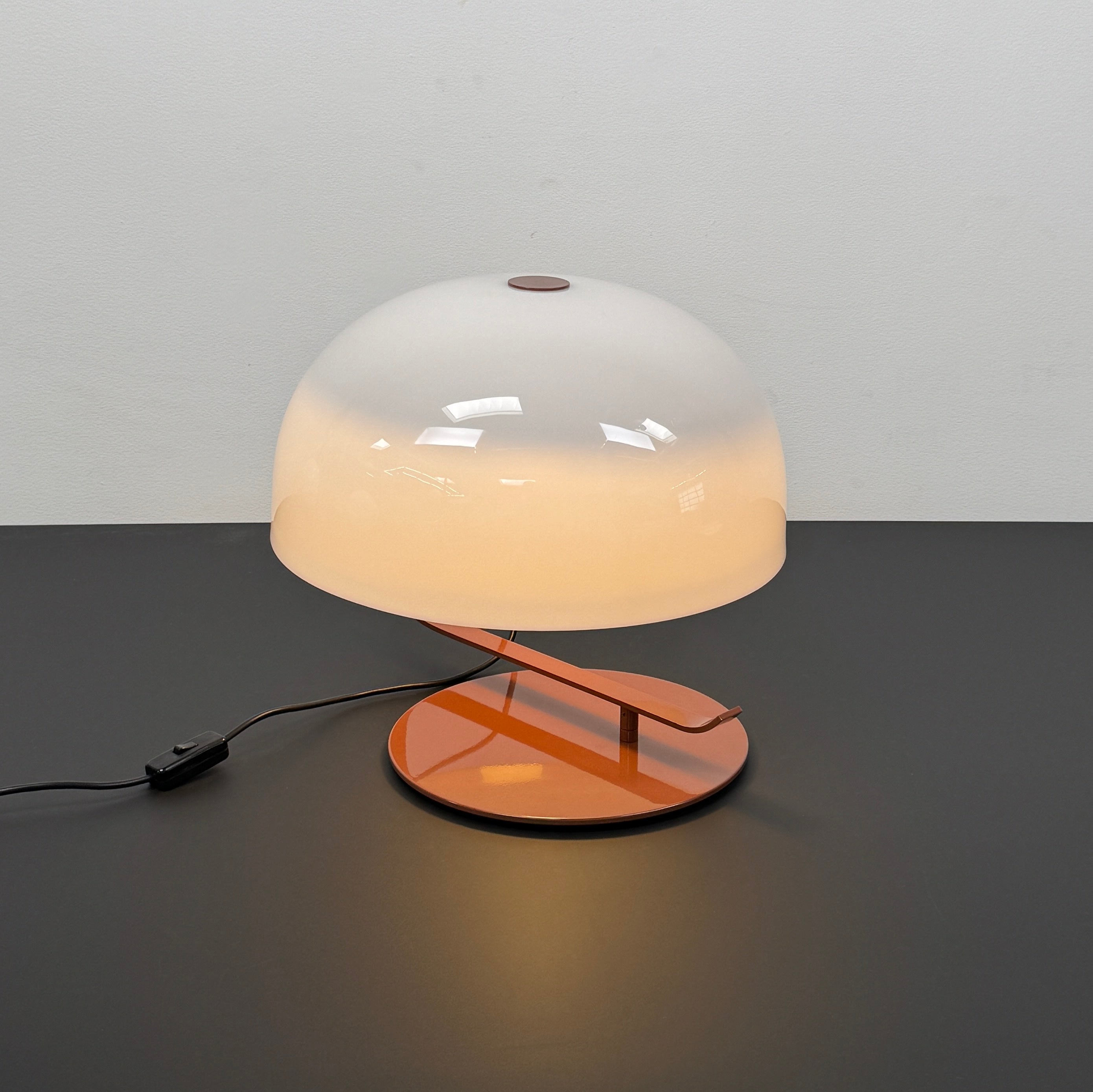 The Oluce Zanuso 275 Table Light by Oluce is a modern lamp inspired by Italian design, with a white domed glass shade, orange base, and black power cord with switch, shown against a plain background.