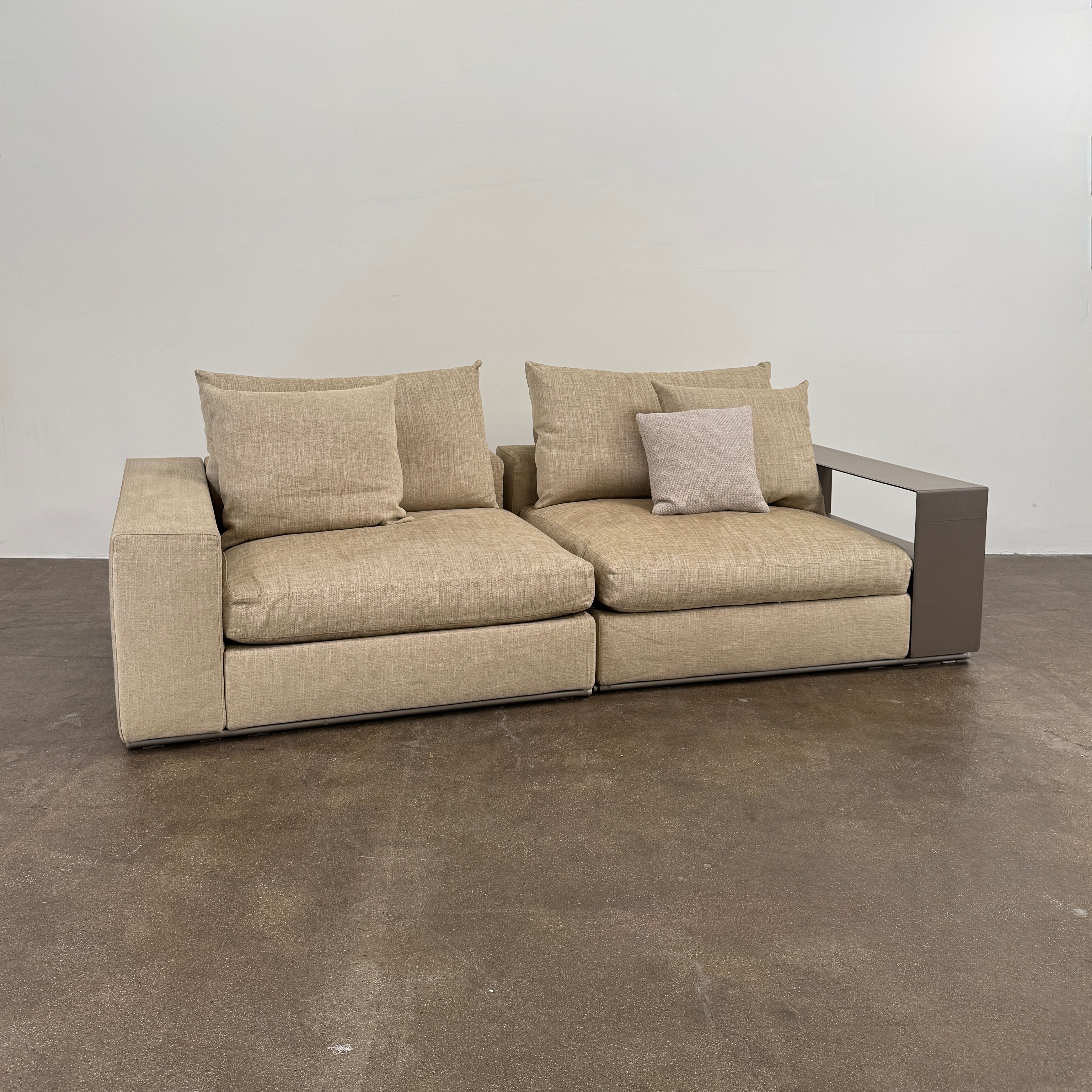 The Flexform Groundpiece Sofa With Leather Shelving by Flexform is a beige two-seater featuring four large cushions, one small beige cushion, and a dark leather shelf attached to the right armrest for sleek modern seating.