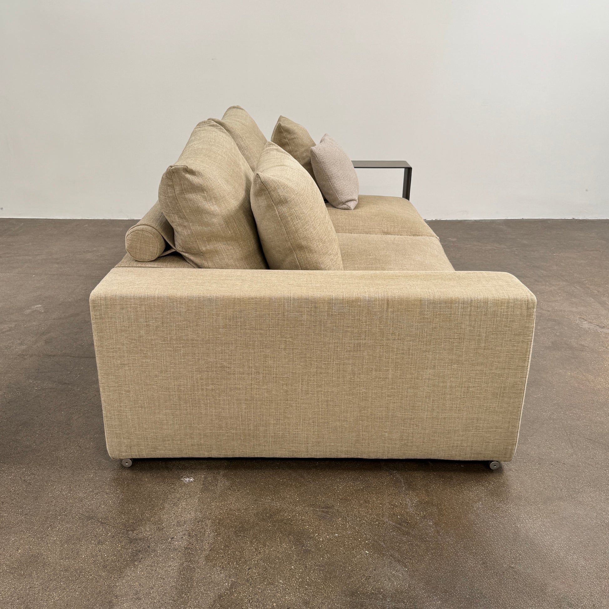 The Flexform Groundpiece Sofa With Leather Shelving by Flexform is a beige two-seater featuring four large cushions, one small beige cushion, and a dark leather shelf attached to the right armrest for sleek modern seating.