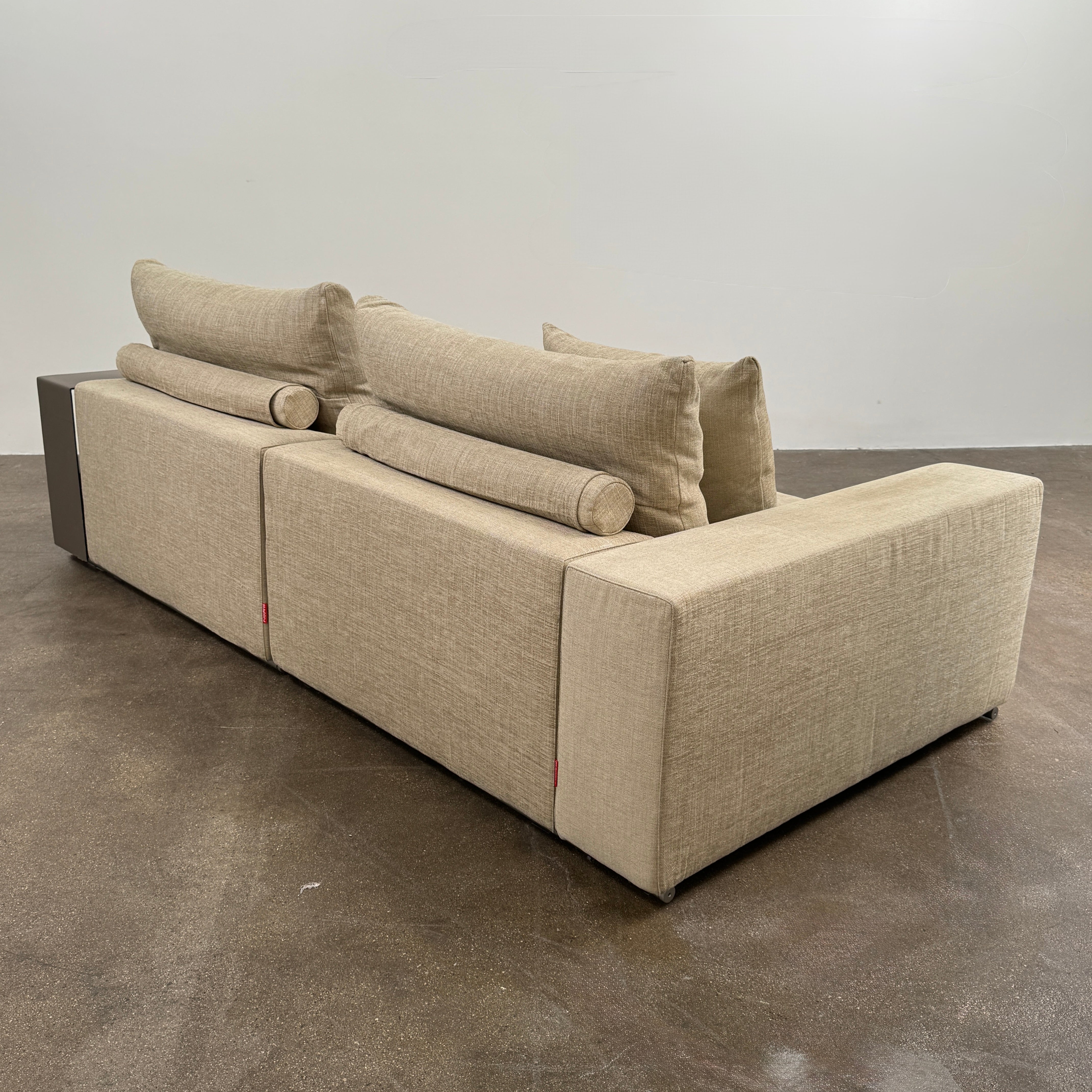 The Flexform Groundpiece Sofa With Leather Shelving by Flexform is a beige two-seater featuring four large cushions, one small beige cushion, and a dark leather shelf attached to the right armrest for sleek modern seating.
