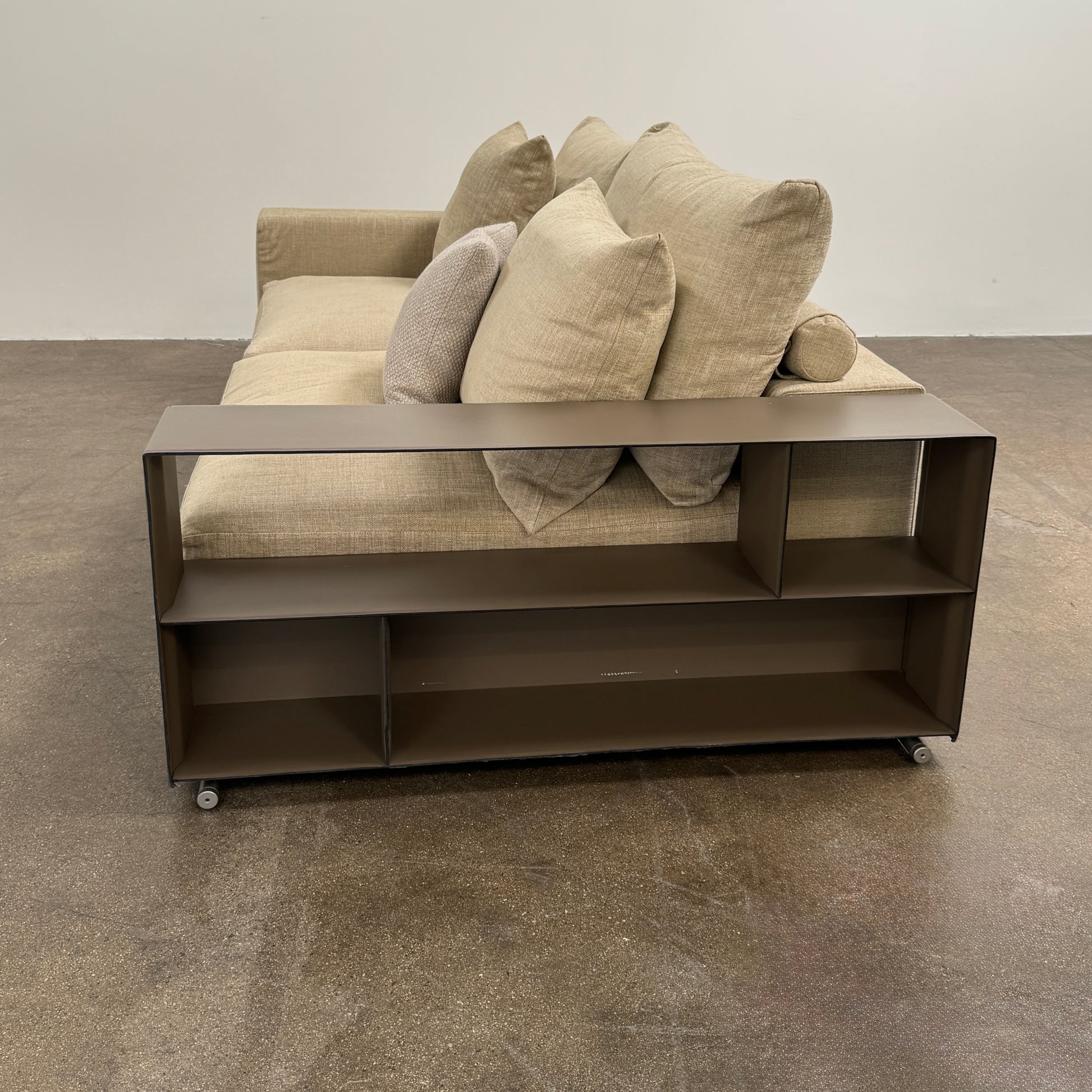 The Flexform Groundpiece Sofa With Leather Shelving by Flexform is a beige two-seater featuring four large cushions, one small beige cushion, and a dark leather shelf attached to the right armrest for sleek modern seating.