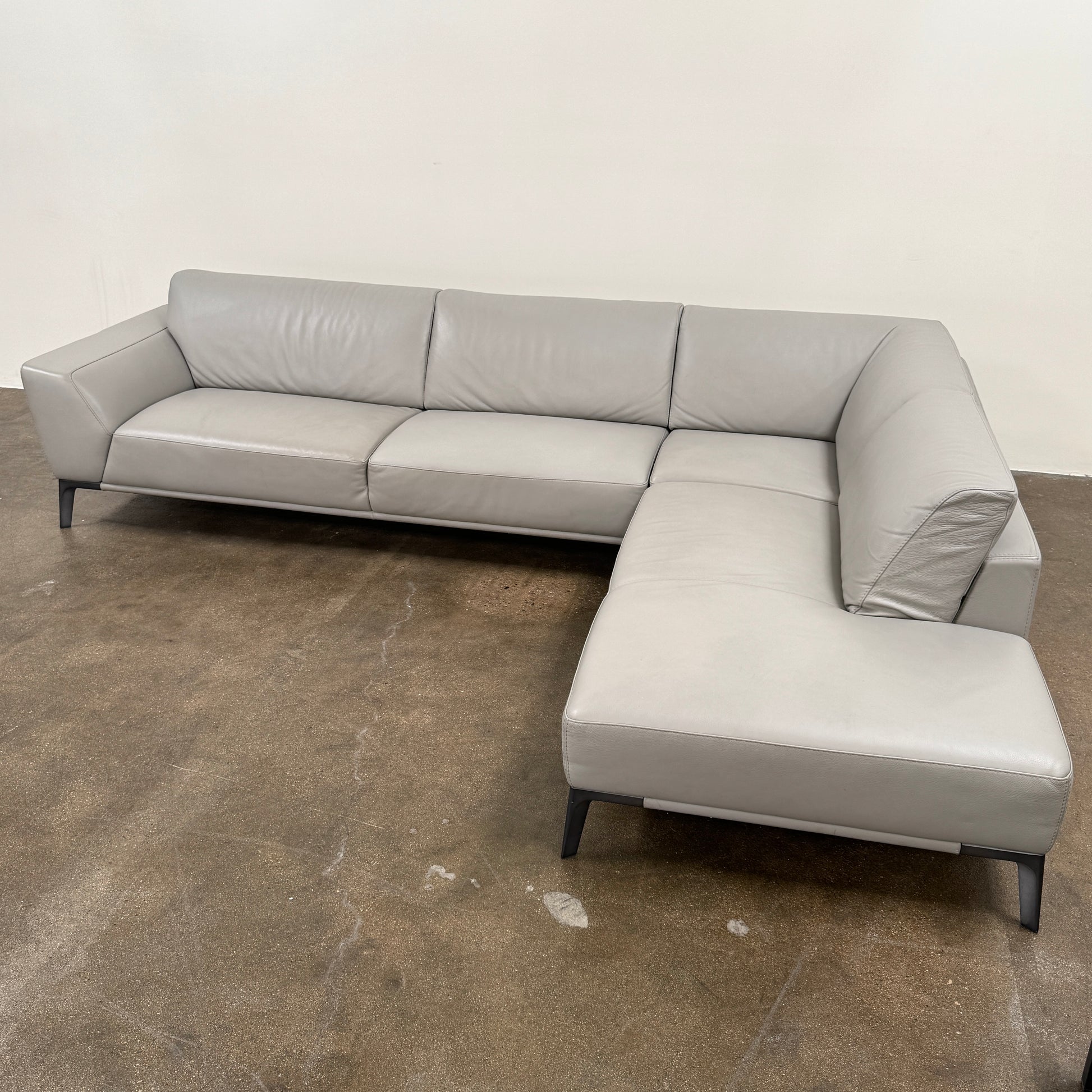A Roche Bobois Accord Gray Leather Sectional sits in a spacious room with floor-to-ceiling windows and light wood floors, overlooking a cityscape.