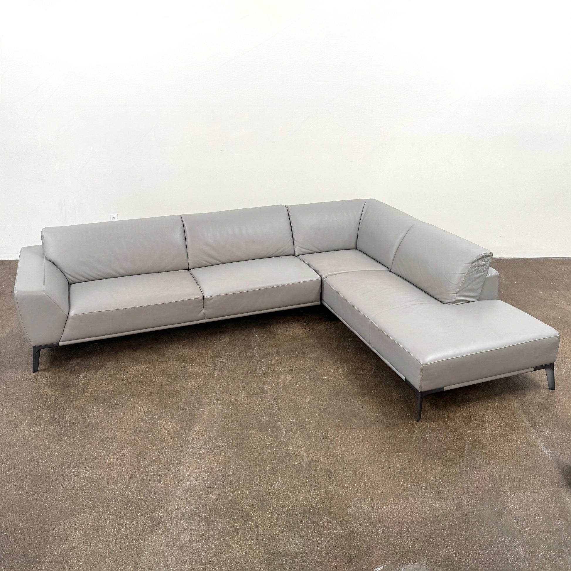 A Roche Bobois Accord Gray Leather Sectional sits in a spacious room with floor-to-ceiling windows and light wood floors, overlooking a cityscape.