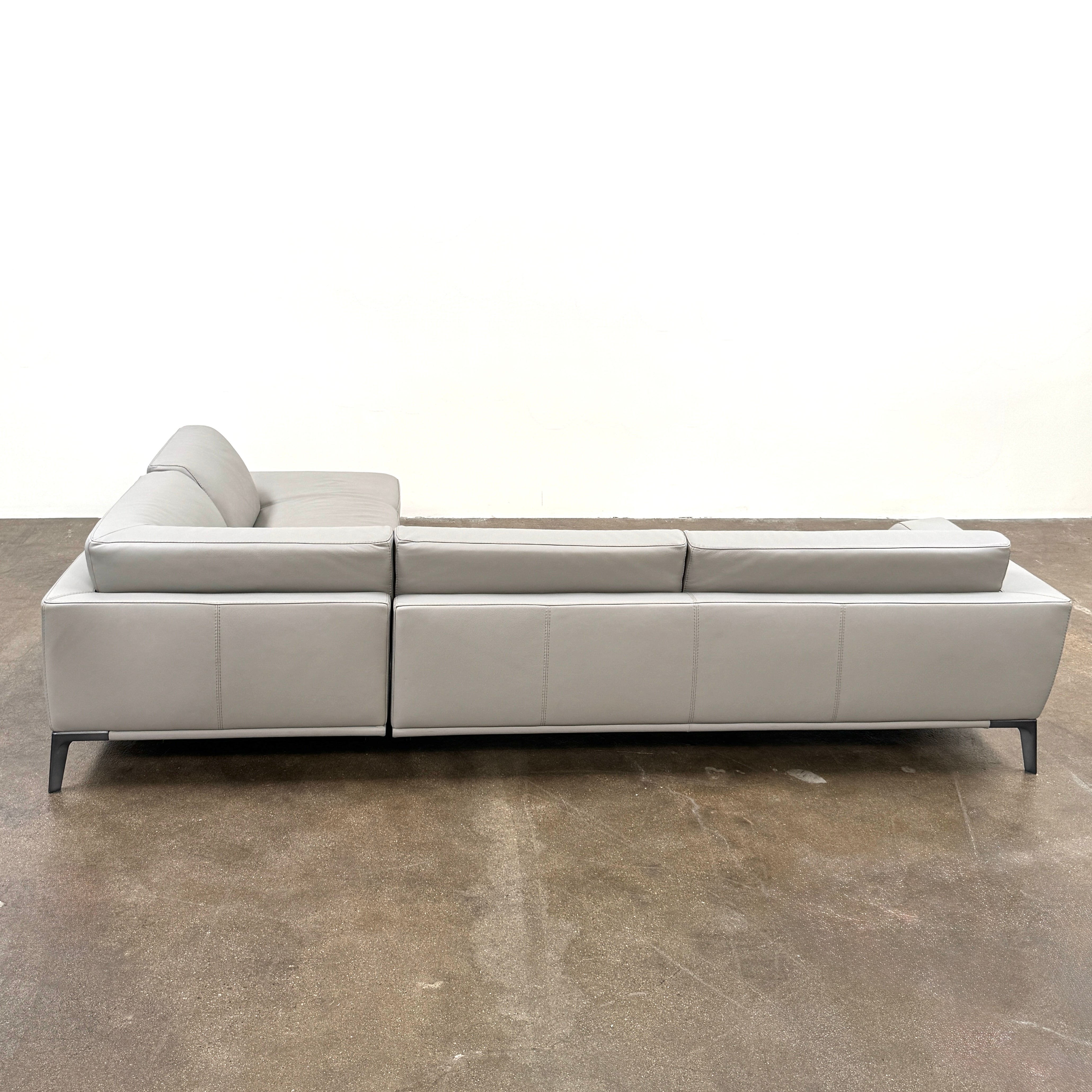 A Roche Bobois Accord Gray Leather Sectional sits in a spacious room with floor-to-ceiling windows and light wood floors, overlooking a cityscape.