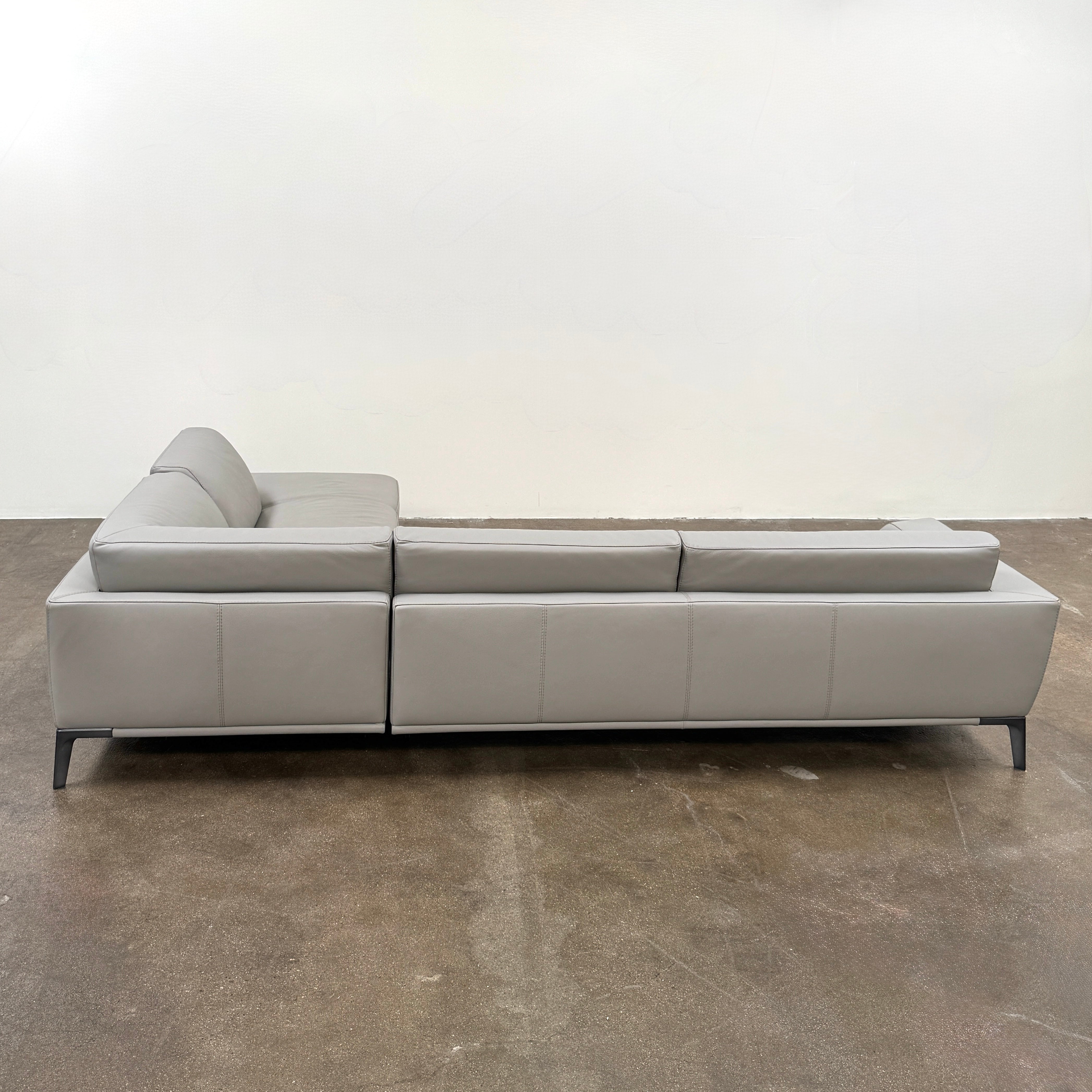 A Roche Bobois Accord Gray Leather Sectional sits in a spacious room with floor-to-ceiling windows and light wood floors, overlooking a cityscape.