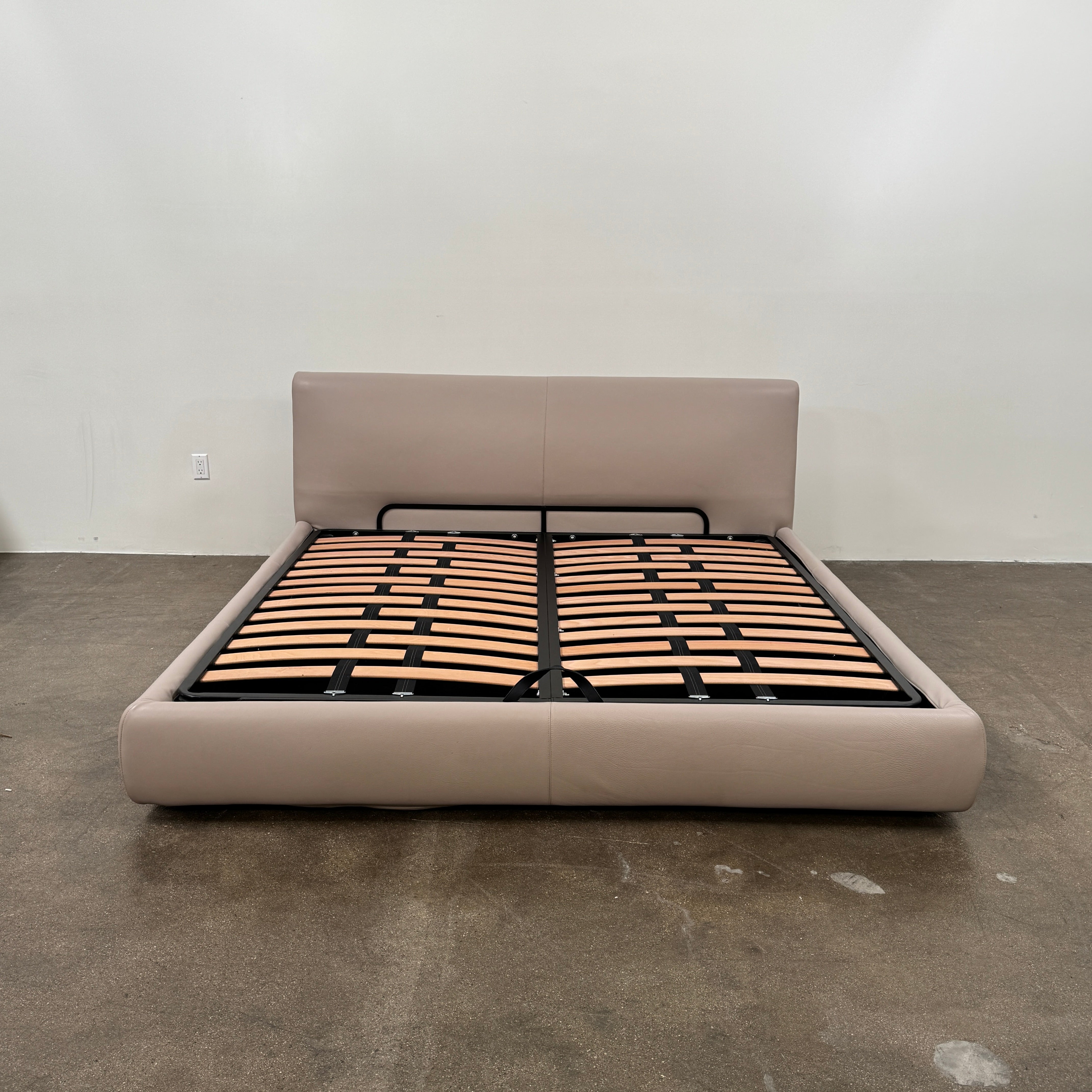 The DWR Nest King Size Leather Bed features a beige upholstered headboard and base, exposed wooden slats, and a sturdy metal support structure for enhanced durability.