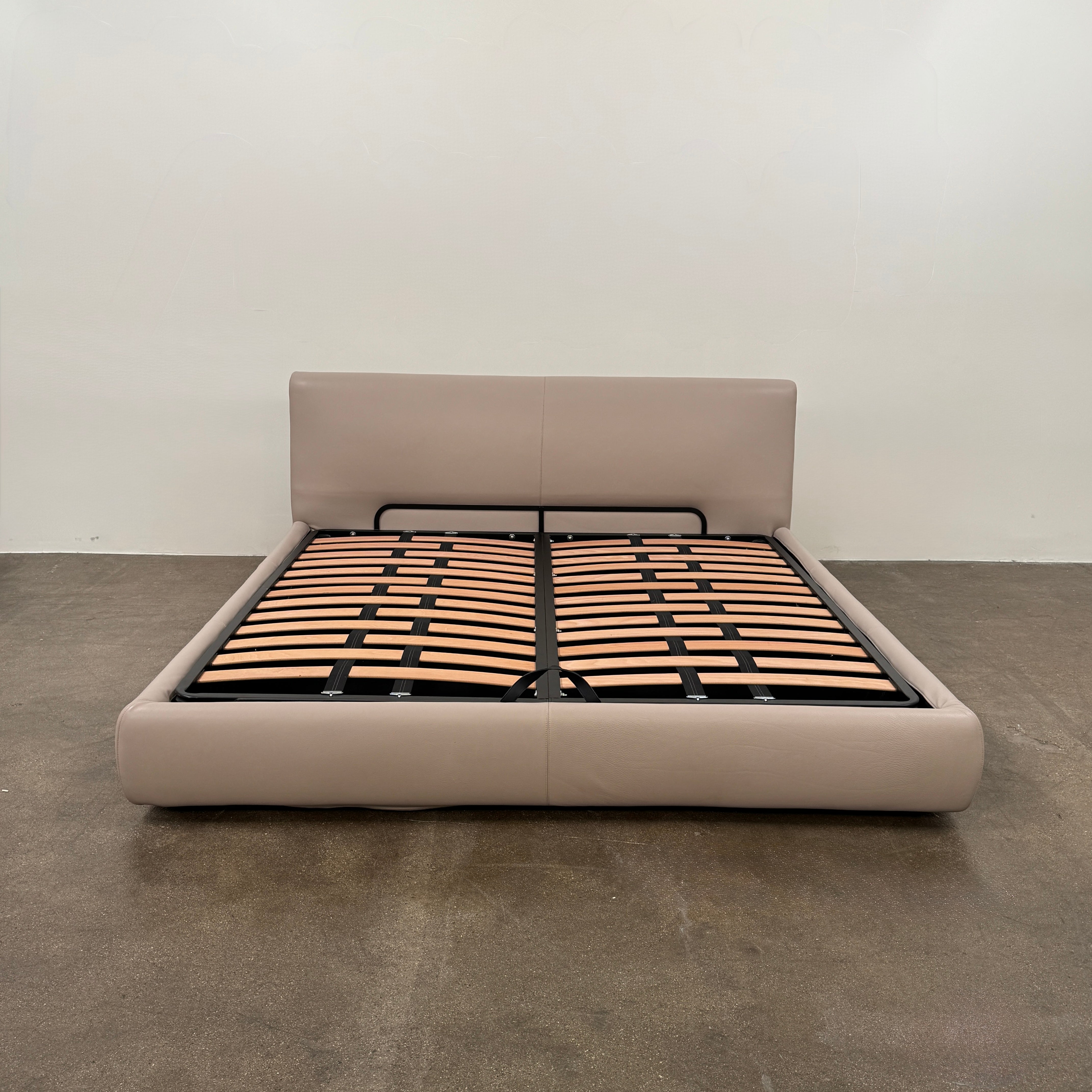 The DWR Nest King Size Leather Bed features a beige upholstered headboard and base, exposed wooden slats, and a sturdy metal support structure for enhanced durability.