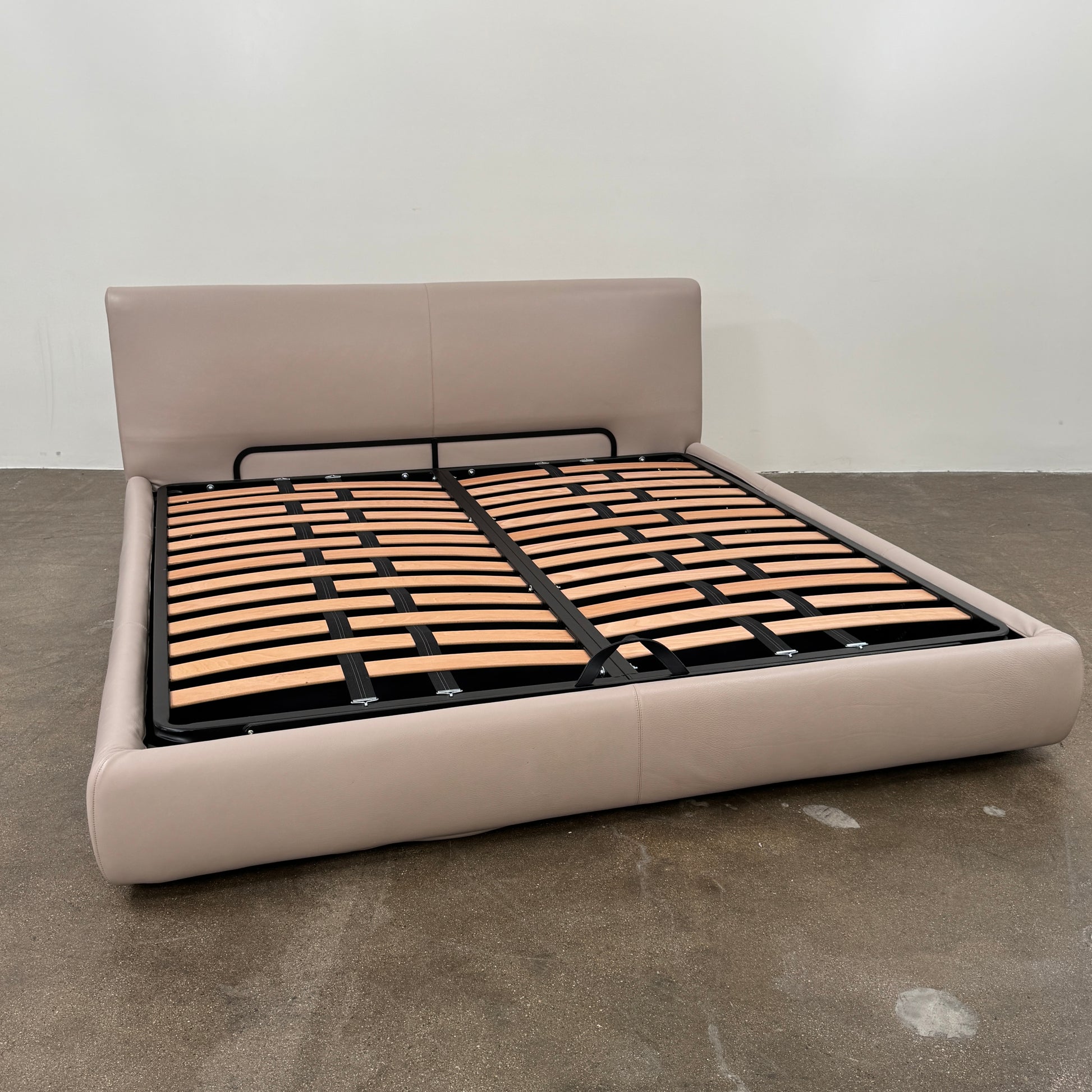 The DWR Nest King Size Leather Bed features a beige upholstered headboard and base, exposed wooden slats, and a sturdy metal support structure for enhanced durability.
