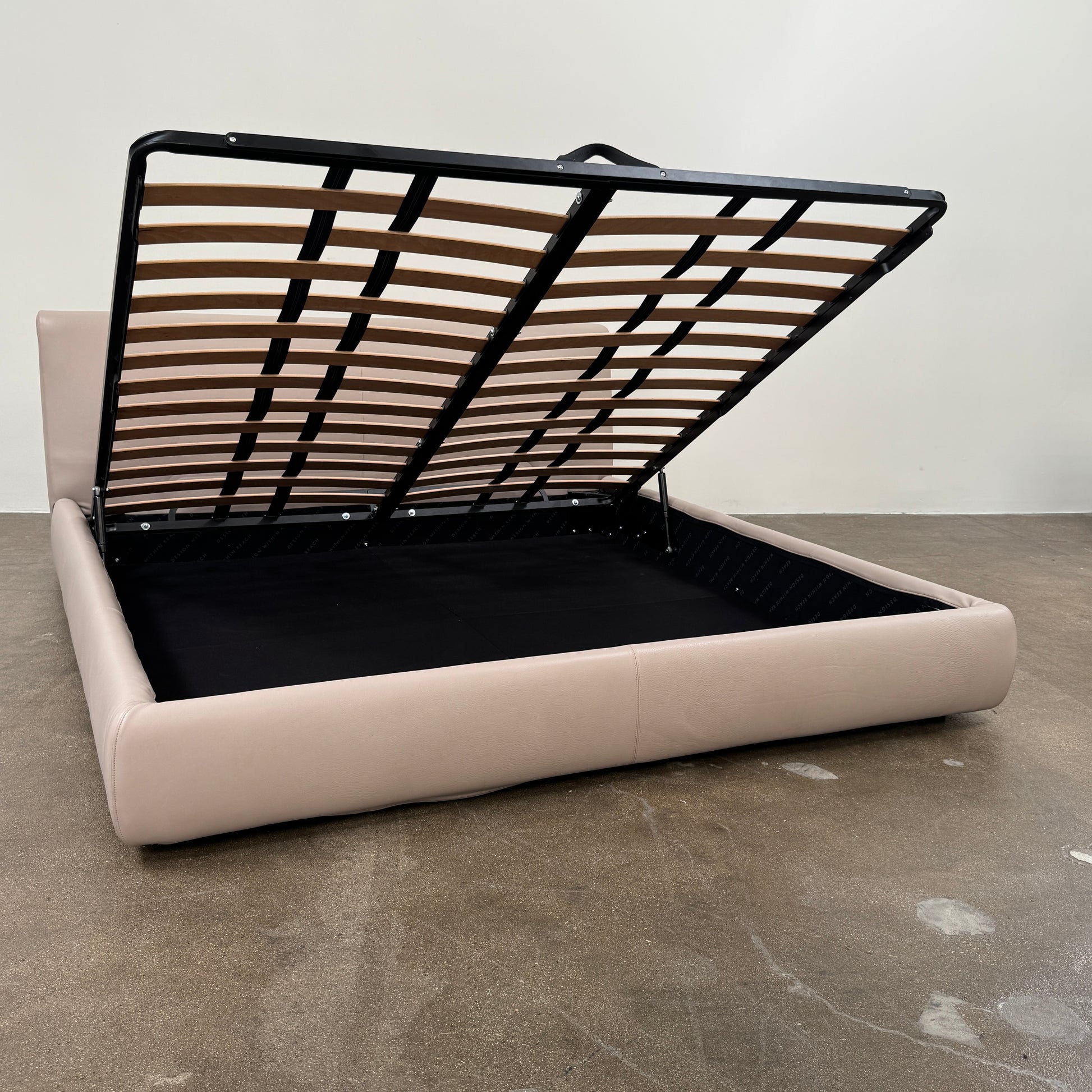 The DWR Nest King Size Leather Bed features a beige upholstered headboard and base, exposed wooden slats, and a sturdy metal support structure for enhanced durability.