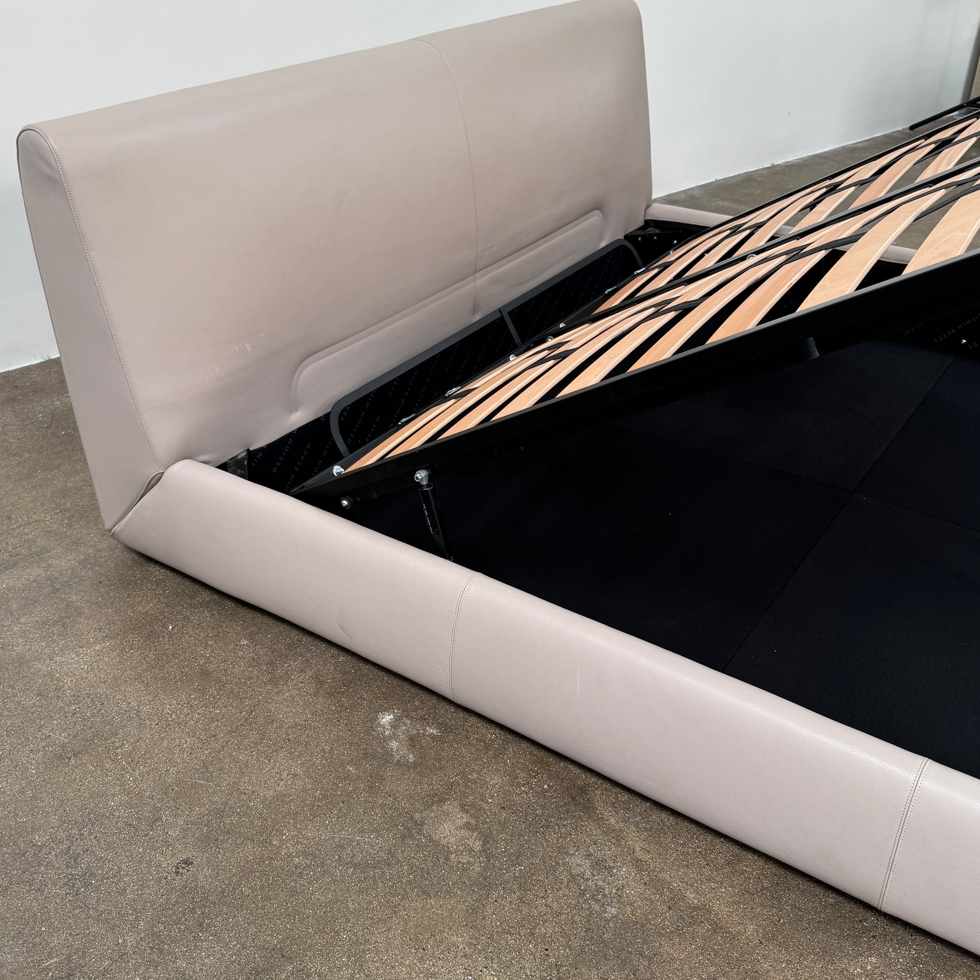 The DWR Nest King Size Leather Bed features a beige upholstered headboard and base, exposed wooden slats, and a sturdy metal support structure for enhanced durability.