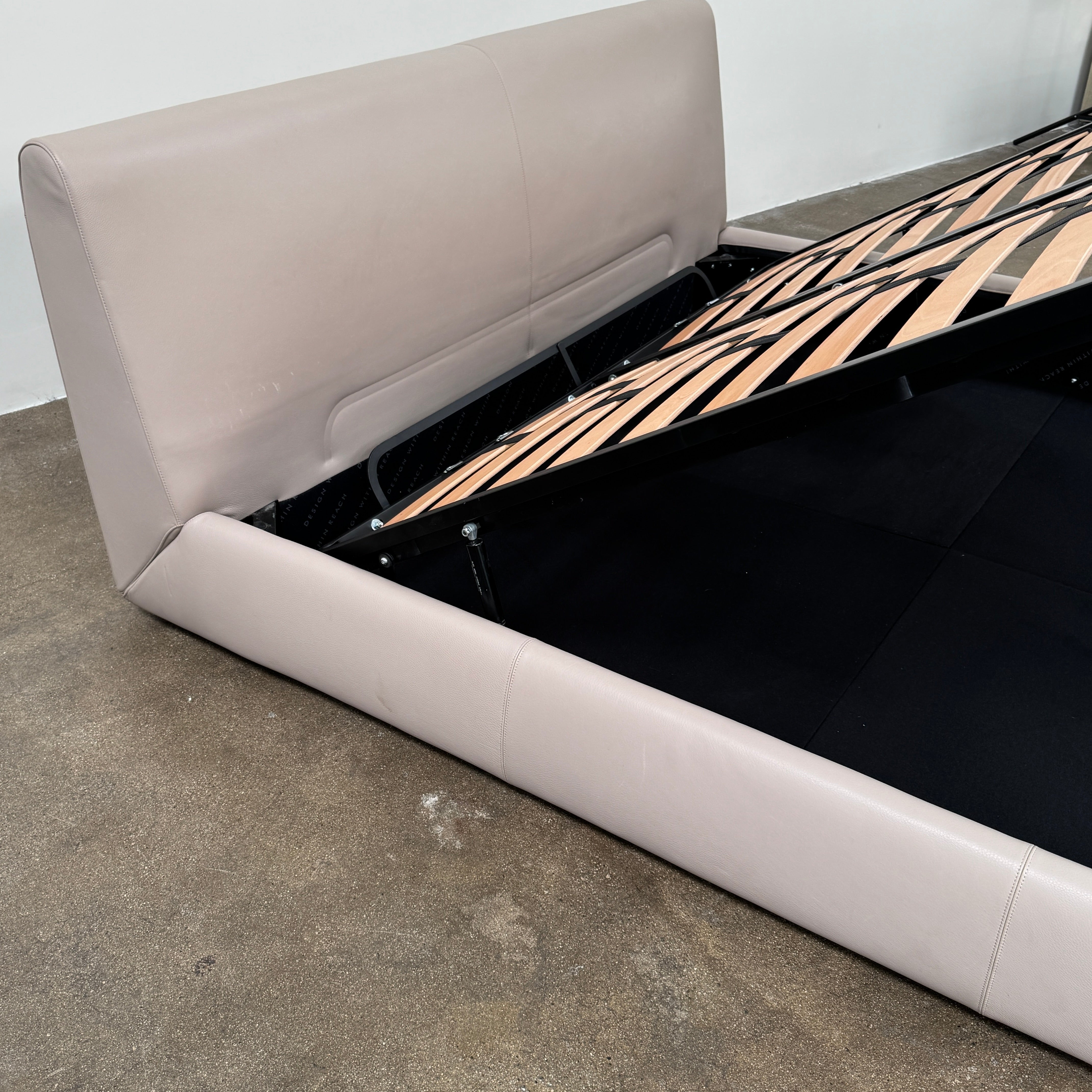 The DWR Nest King Size Leather Bed features a beige upholstered headboard and base, exposed wooden slats, and a sturdy metal support structure for enhanced durability.